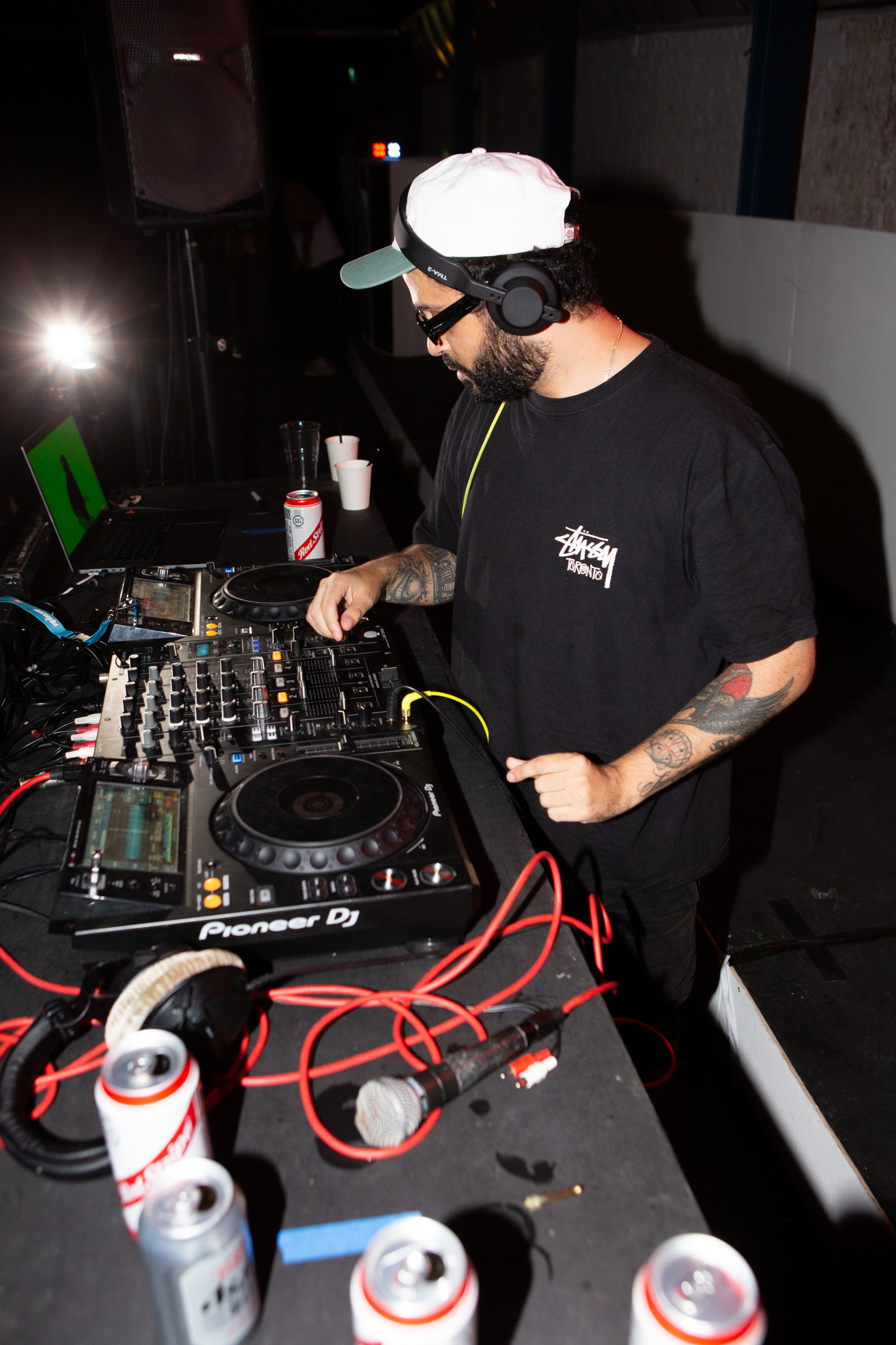 DJ with white cap and headphones operating Pioneer mixing deck in dimly lit venue with red cables and drinks nearby.