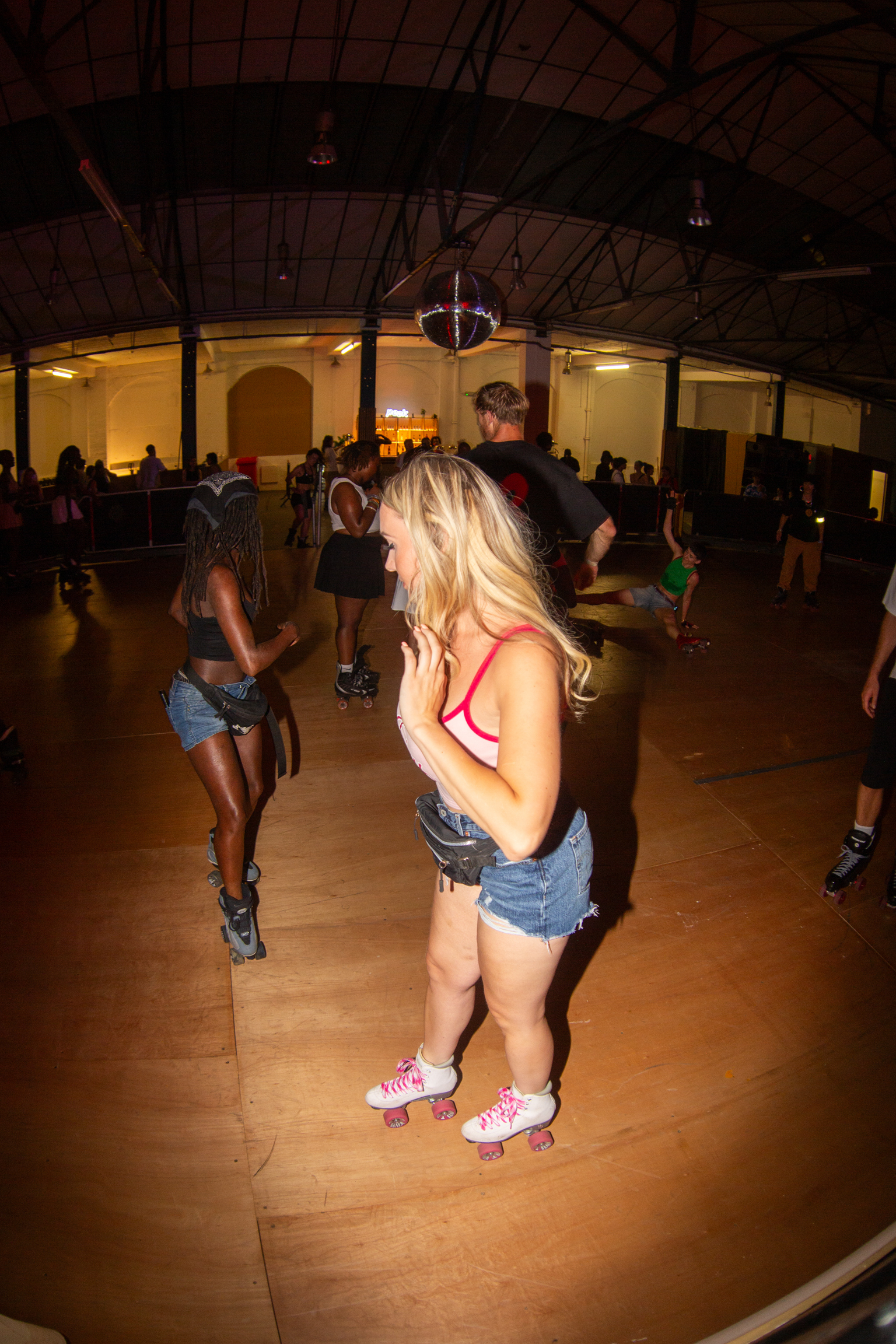 Blonde woman in red bikini top and denim shorts roller skating on wooden rink floor with other skaters in dimly lit venue.