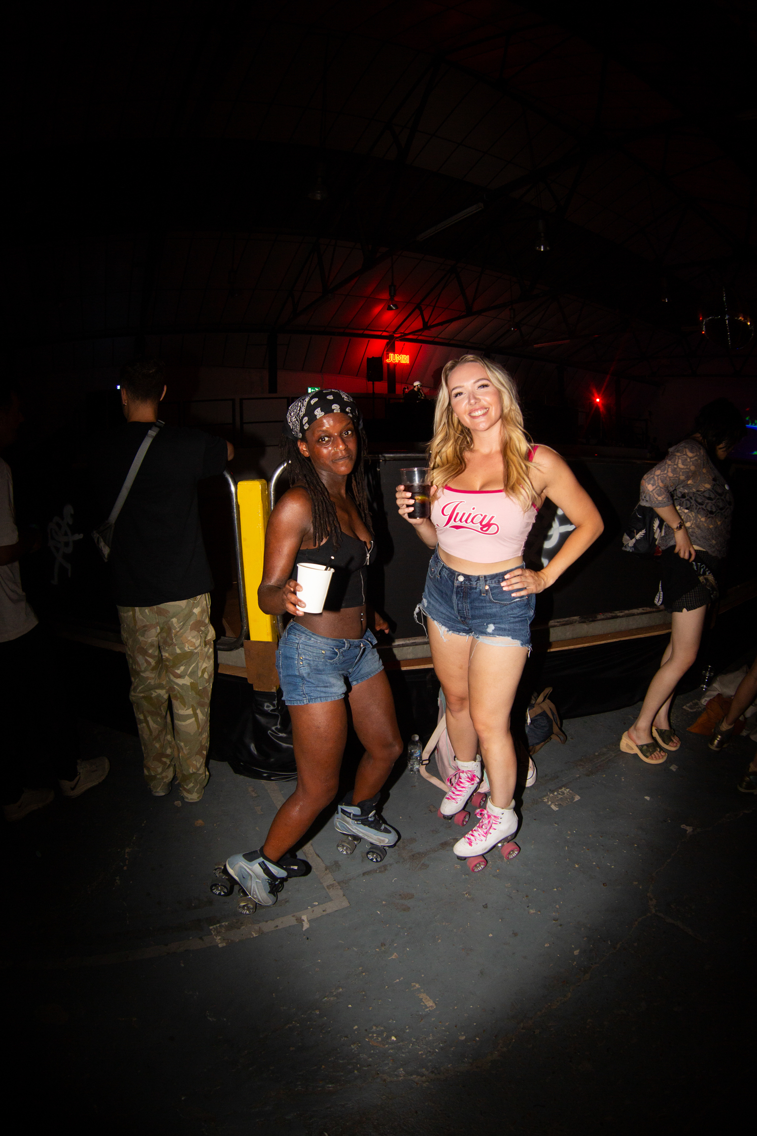 Two women posing with drinks at night club. One wears denim shorts and crop top, other in pink roller skates and "Juicy" top.