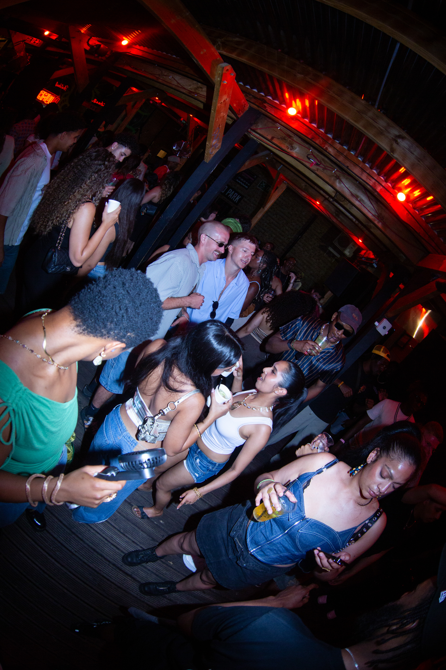 Crowded nightclub scene with people dancing and socialising under red lighting and exposed ceiling beams.