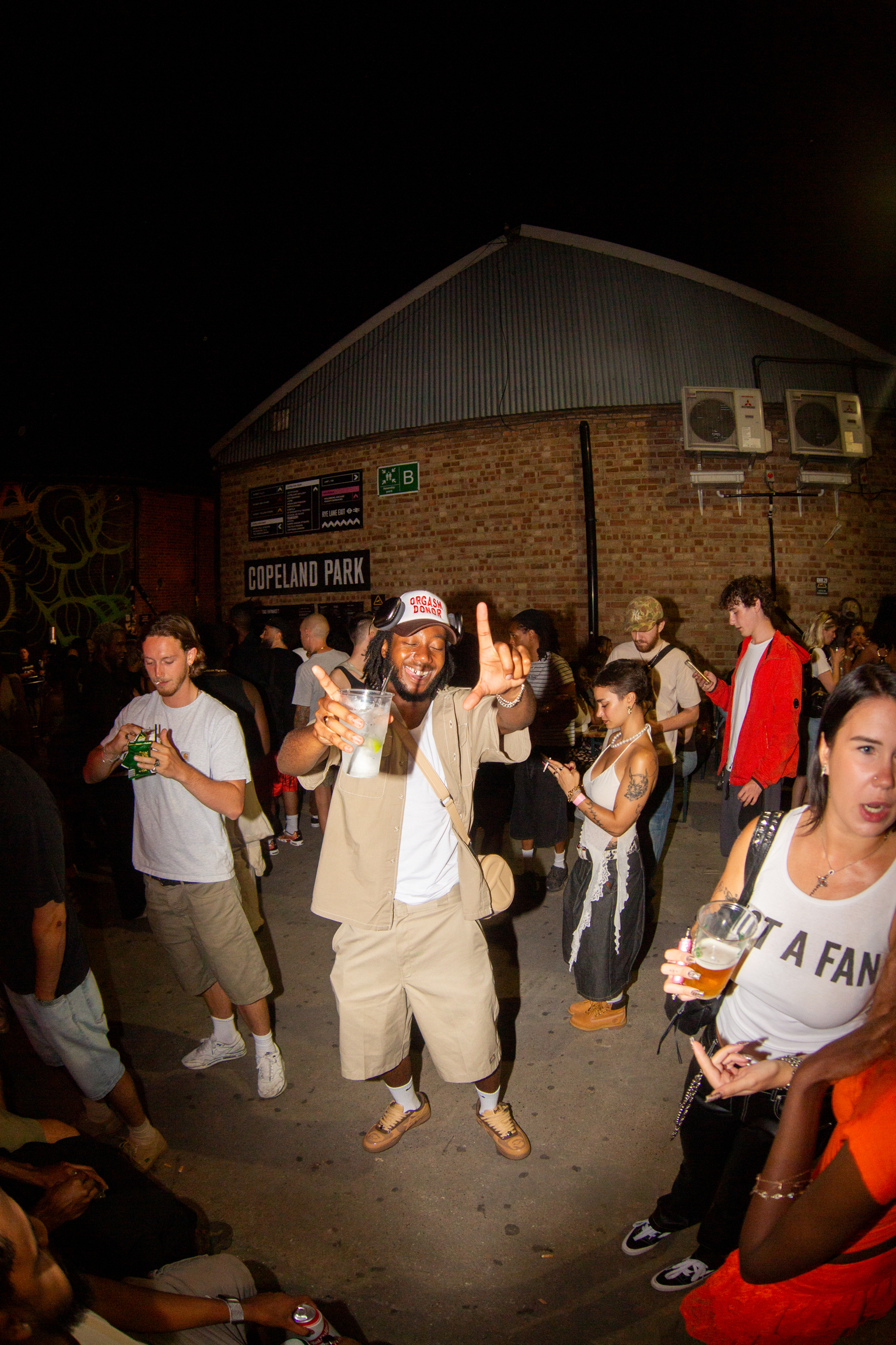 Group of people celebrating at night outside brick building with corrugated roof. Man in centre wearing beige outfit and cap with arms raised.