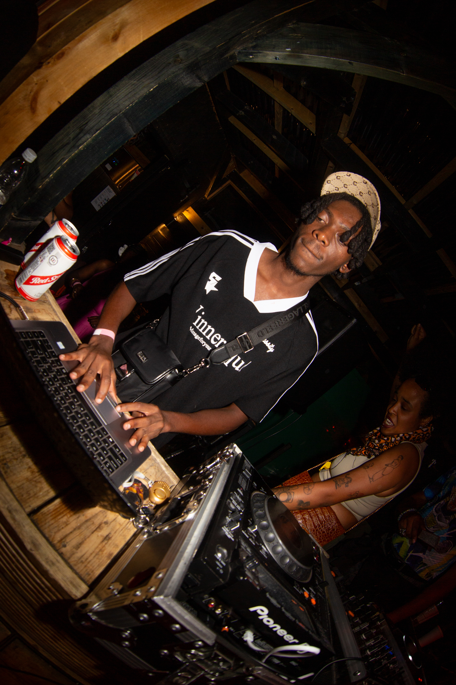DJ wearing black shirt and cap operating mixing equipment in dimly lit venue with wooden beams overhead.