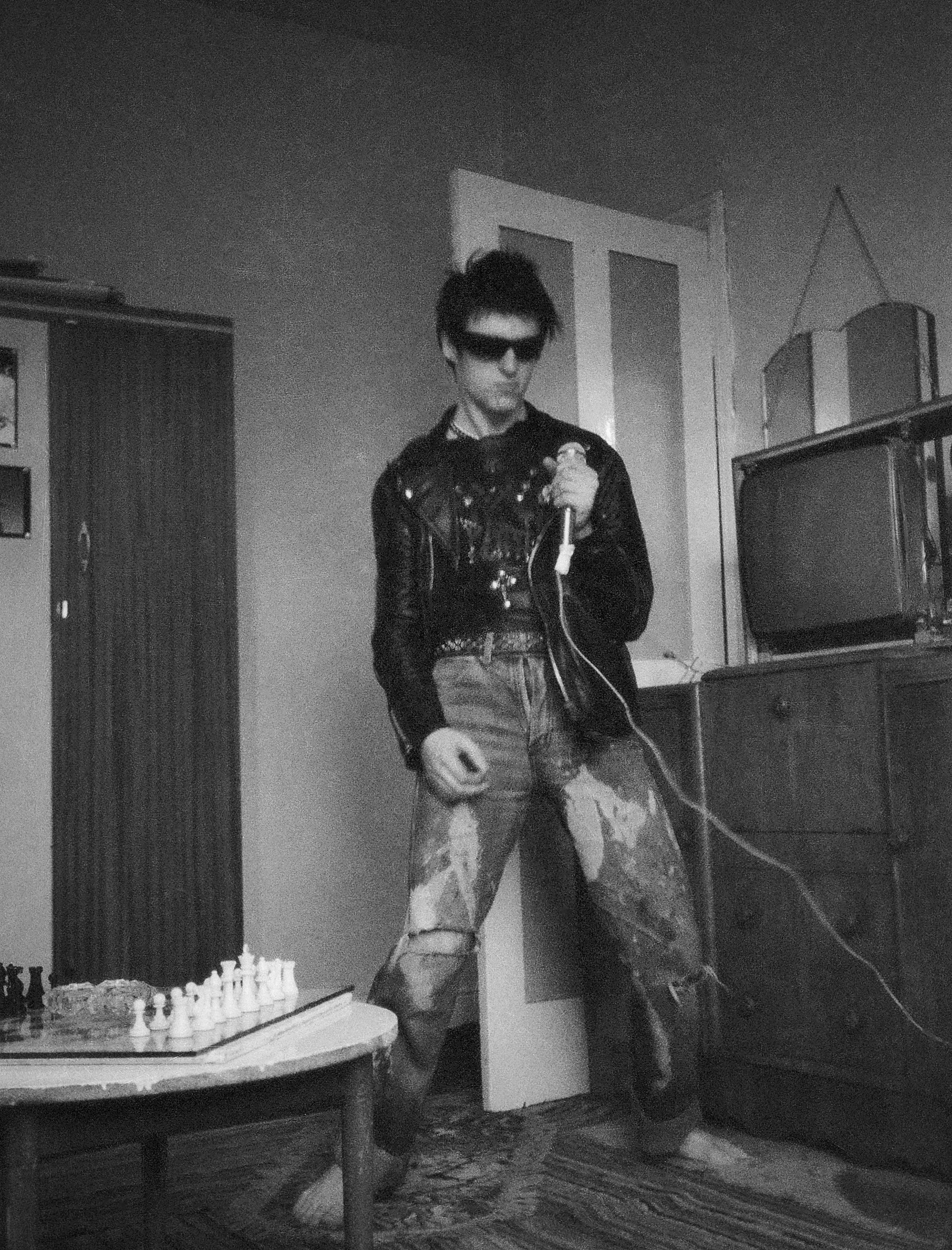 Man in sunglasses and leather jacket holding microphone in domestic interior, black and white photograph with chess set on table.