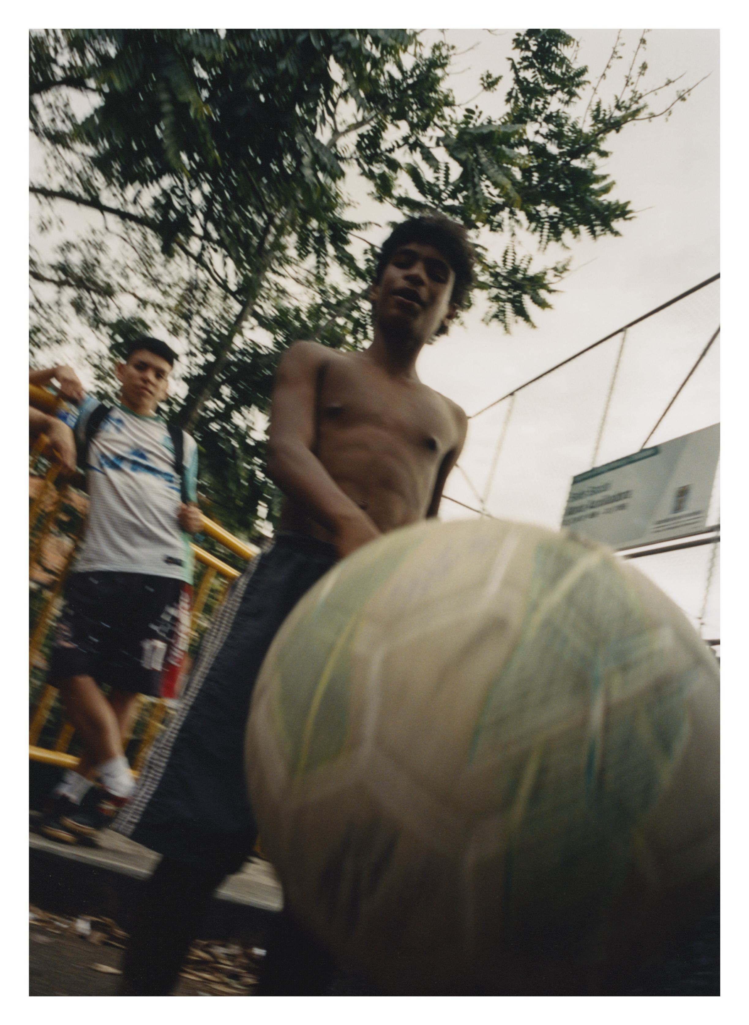 Shirtless man standing behind large pale green spherical object, with people in background near yellow railings and trees overhead.
