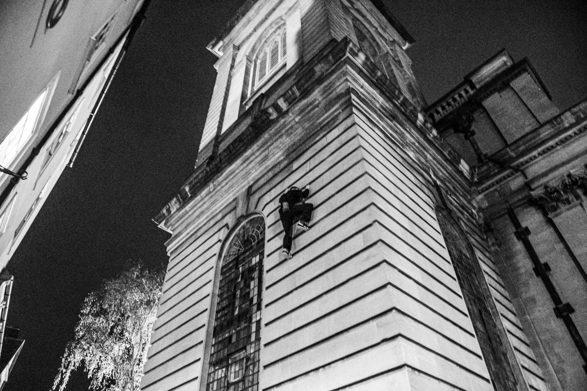 Black and white low-angle view of stone building corner with chevron pattern stonework and arched window against dark sky.