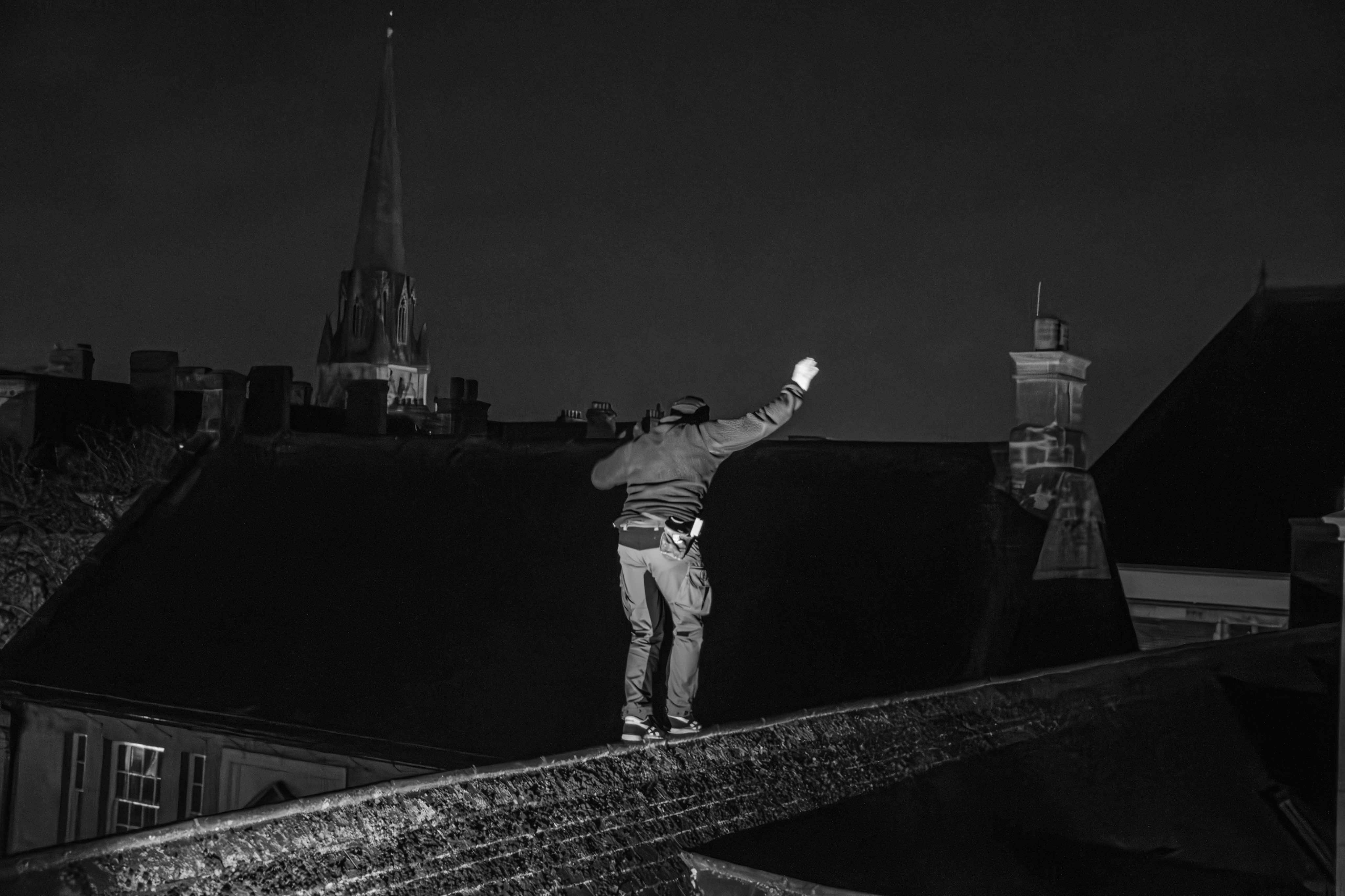 Black and white image: person with arms outstretched walking on narrow ledge or wall, city buildings and spire visible in background