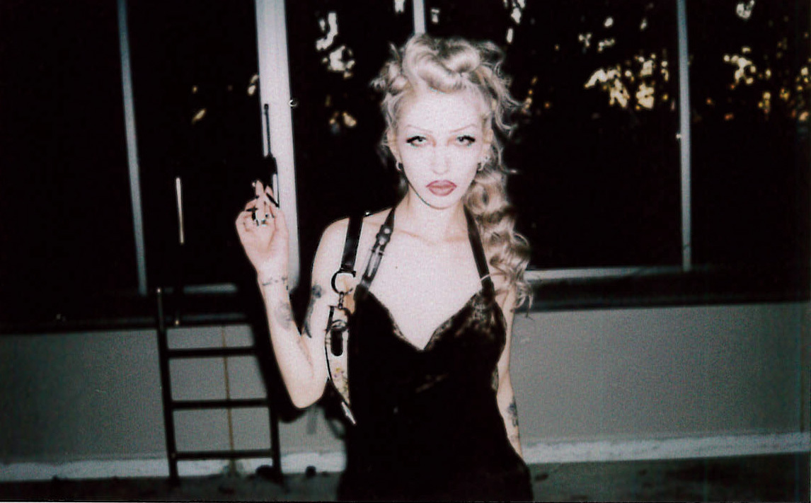 A woman with curly blonde hair wearing a black dress and making a peace sign gesture in front of a dark background.