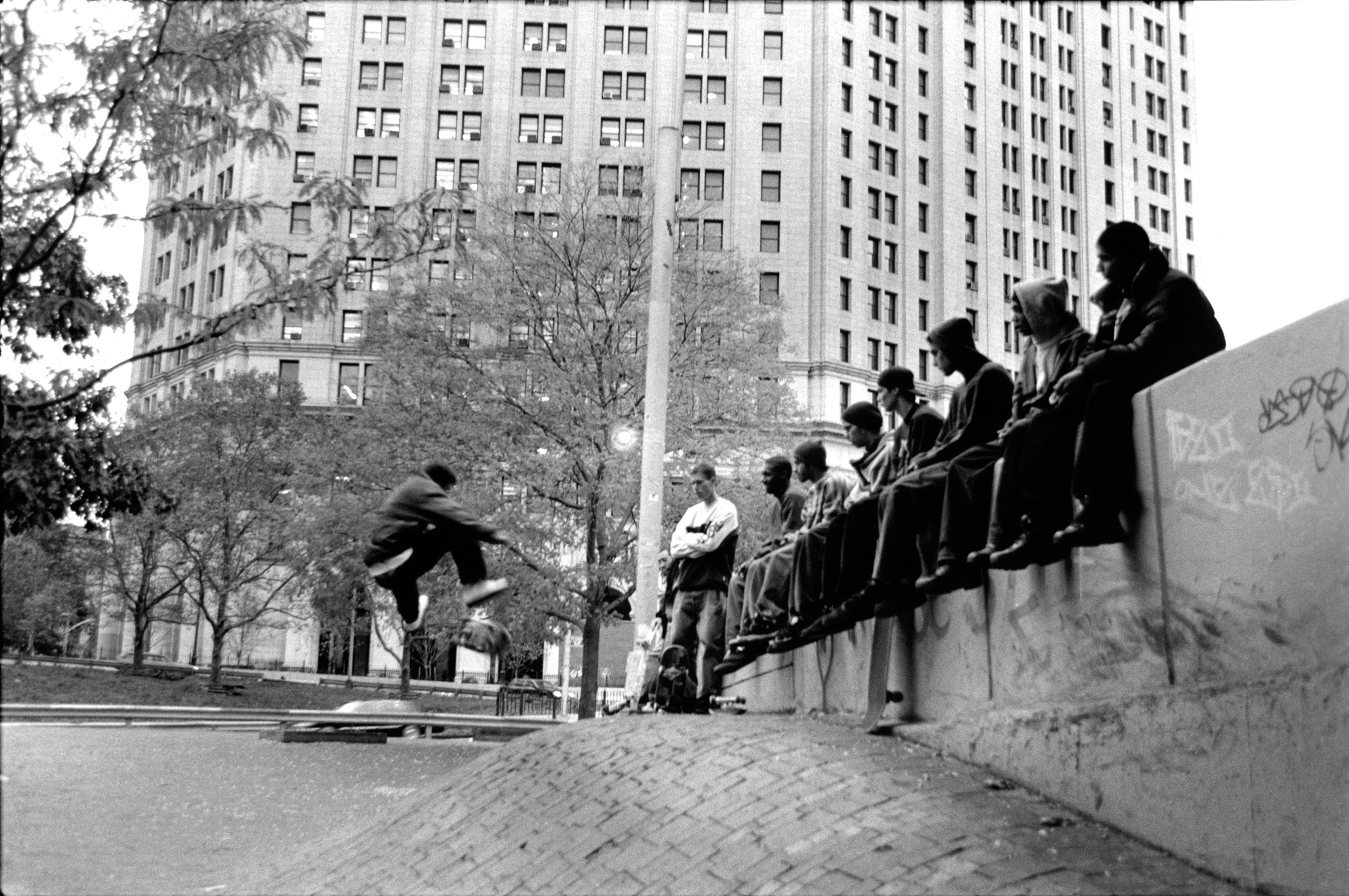 Black and white image: skateboarder mid-air over concrete bowl, group watching from ledge, tall buildings and bare trees in background.