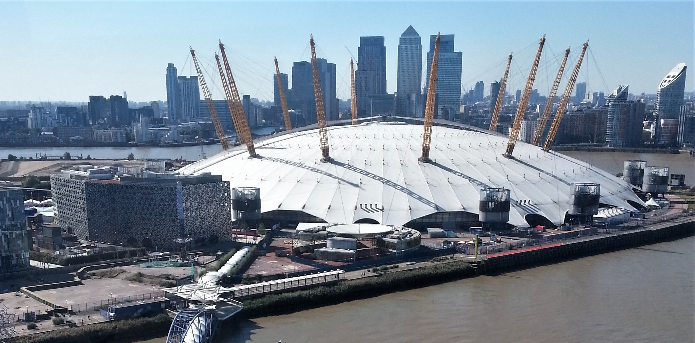 Large white domed arena with yellow support masts beside River Thames, with Canary Wharf skyscrapers in background under blue sky.