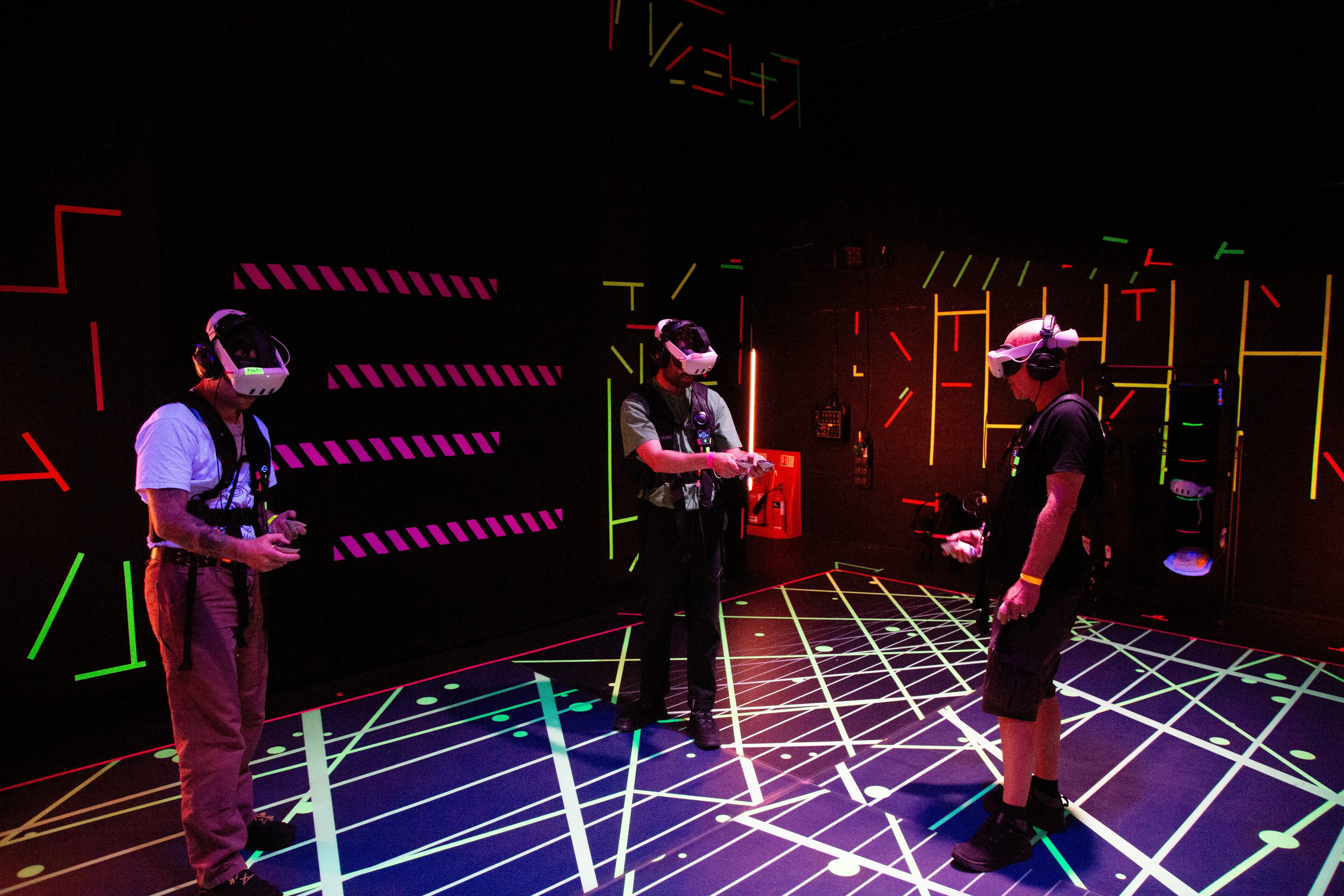 Three people wearing VR headsets in darkened room with neon geometric patterns on walls and floor in pink, green, and blue colours.