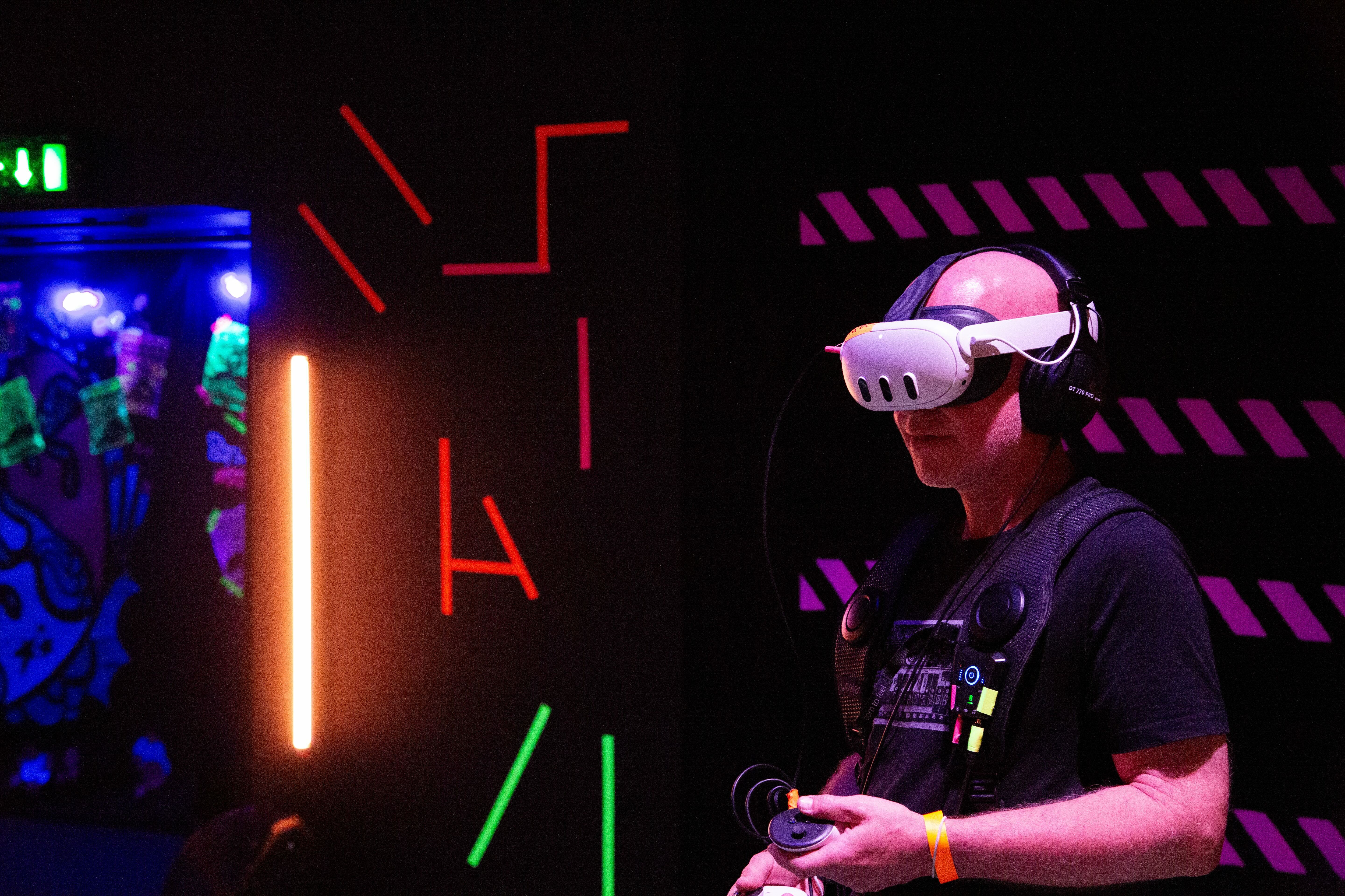Person wearing white VR headset in dark room with neon lighting - blue, red, green, and purple colours illuminating walls and surfaces.