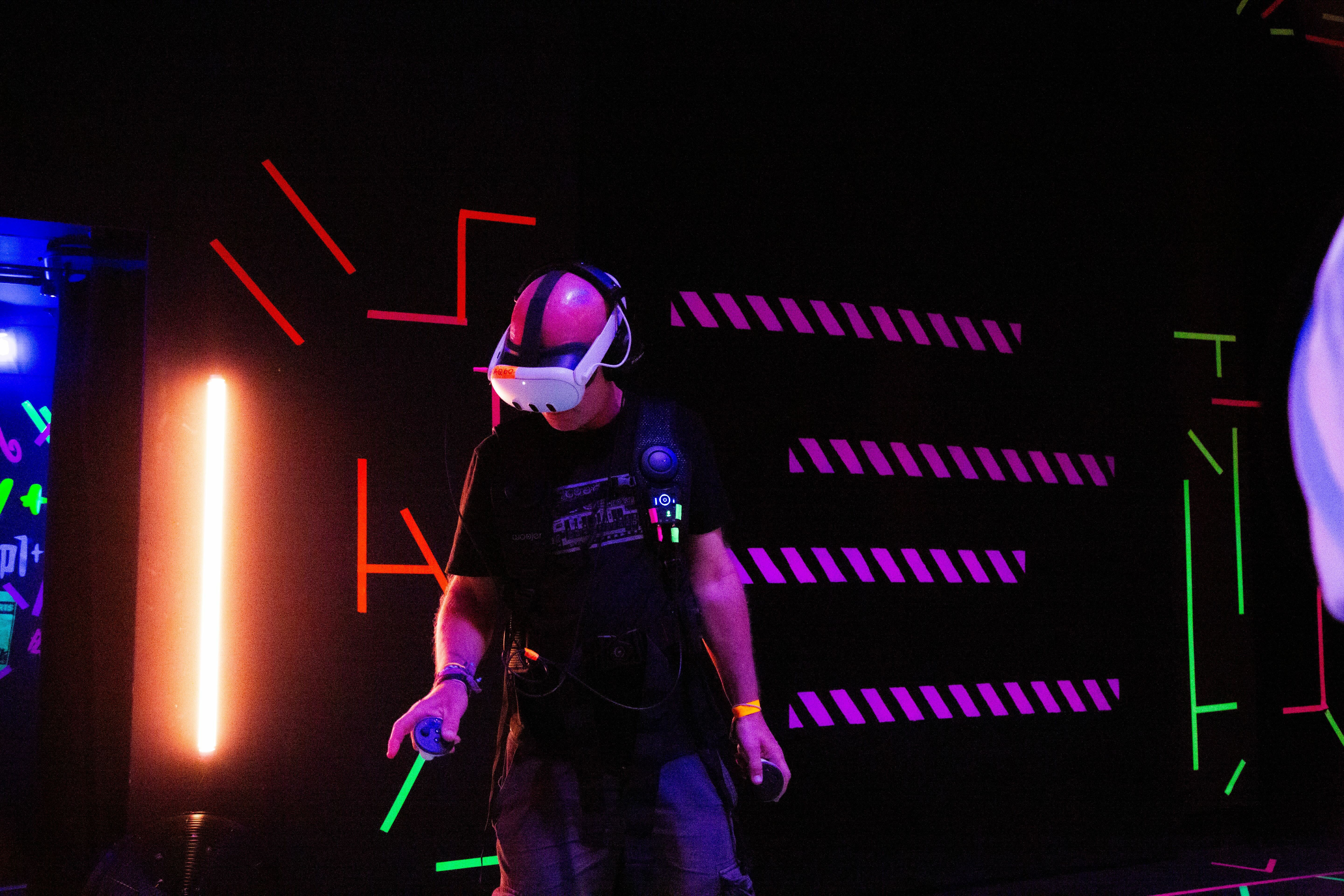 Person in futuristic helmet and dark clothing surrounded by neon lights - orange, purple, green, and blue geometric patterns against black background.