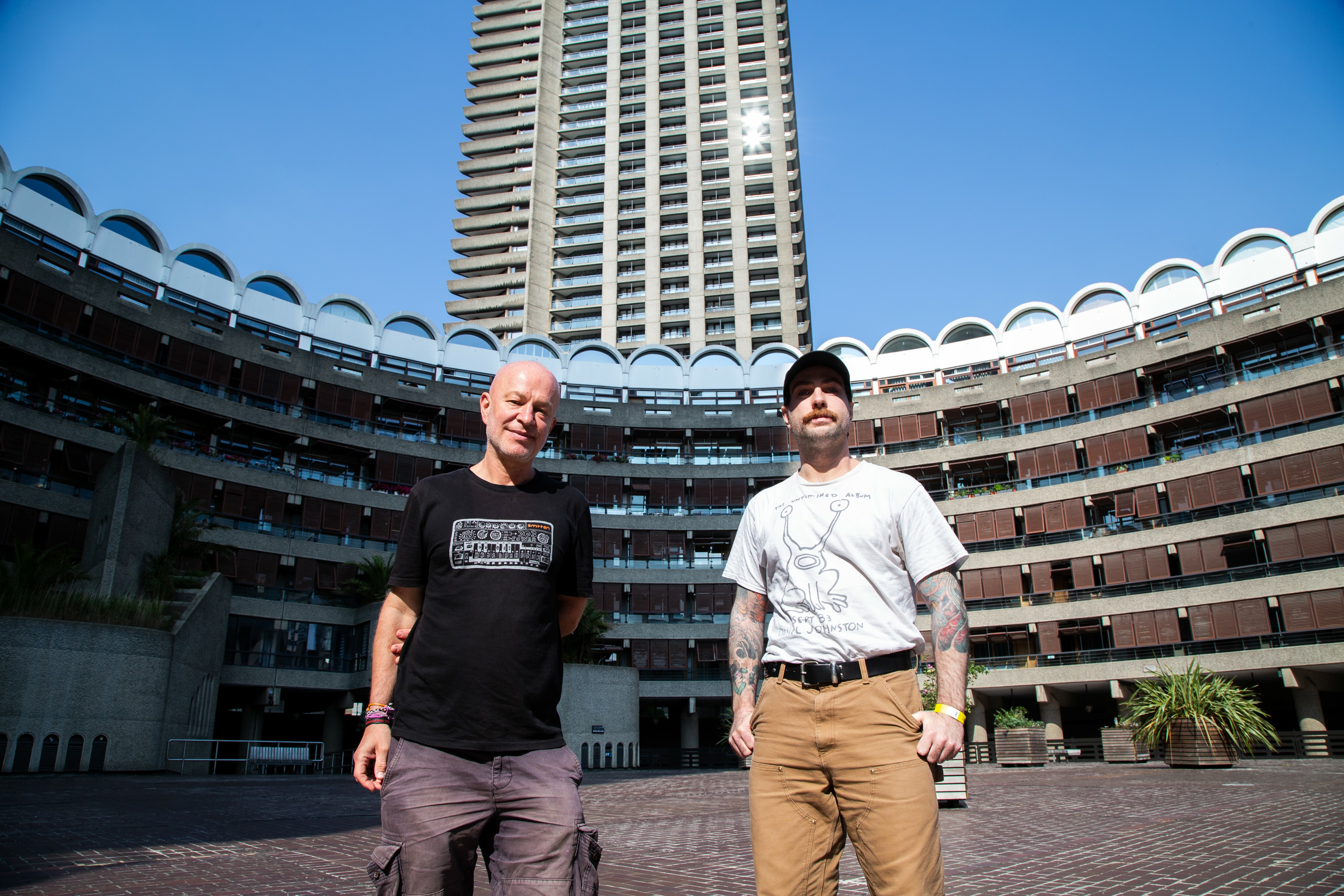 Two men standing in courtyard surrounded by concrete residential buildings with white arched balconies and tall tower block behind.