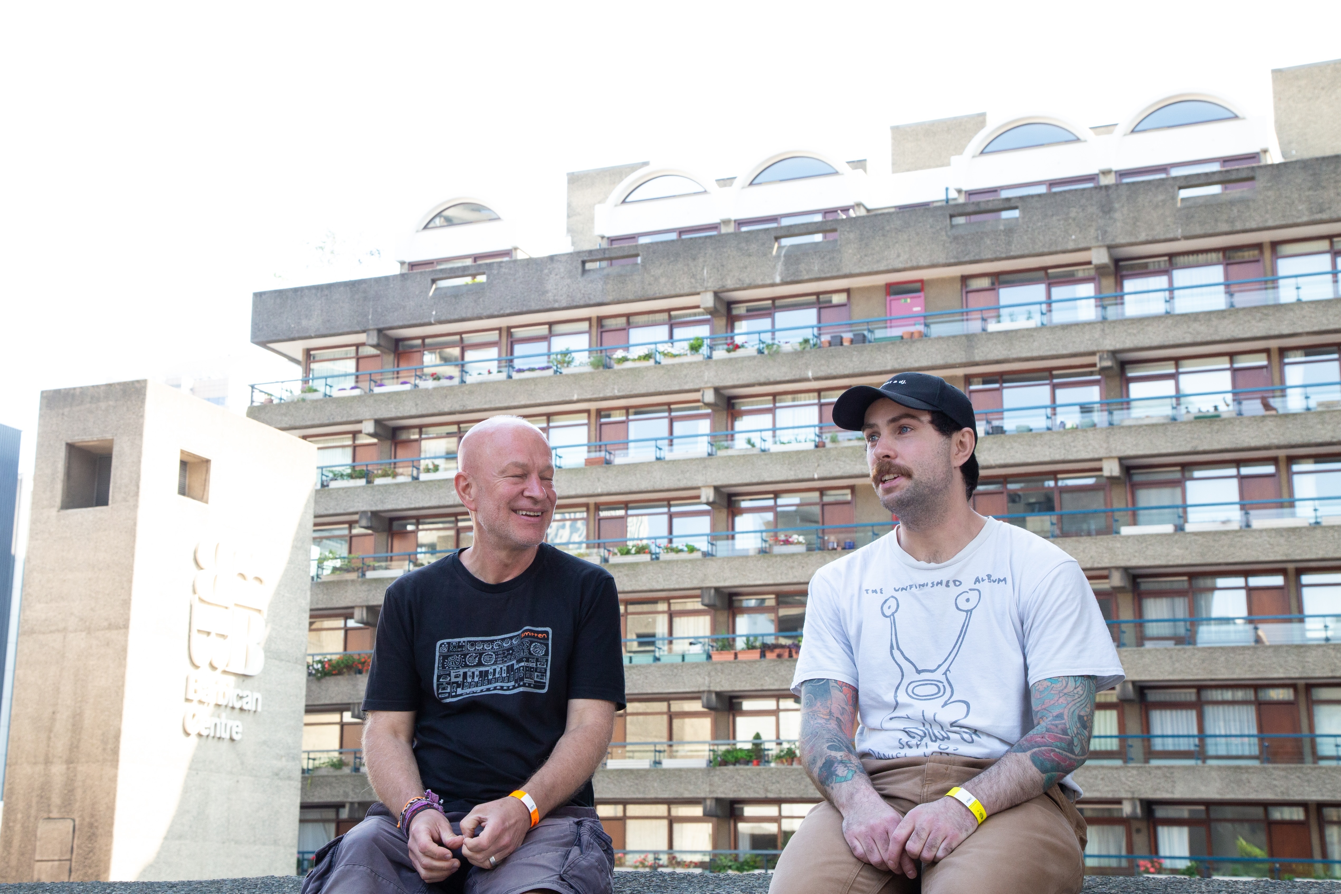 Two men sitting outdoors with multi-storey residential building in background. One wears black t-shirt, other in white with baseball cap.