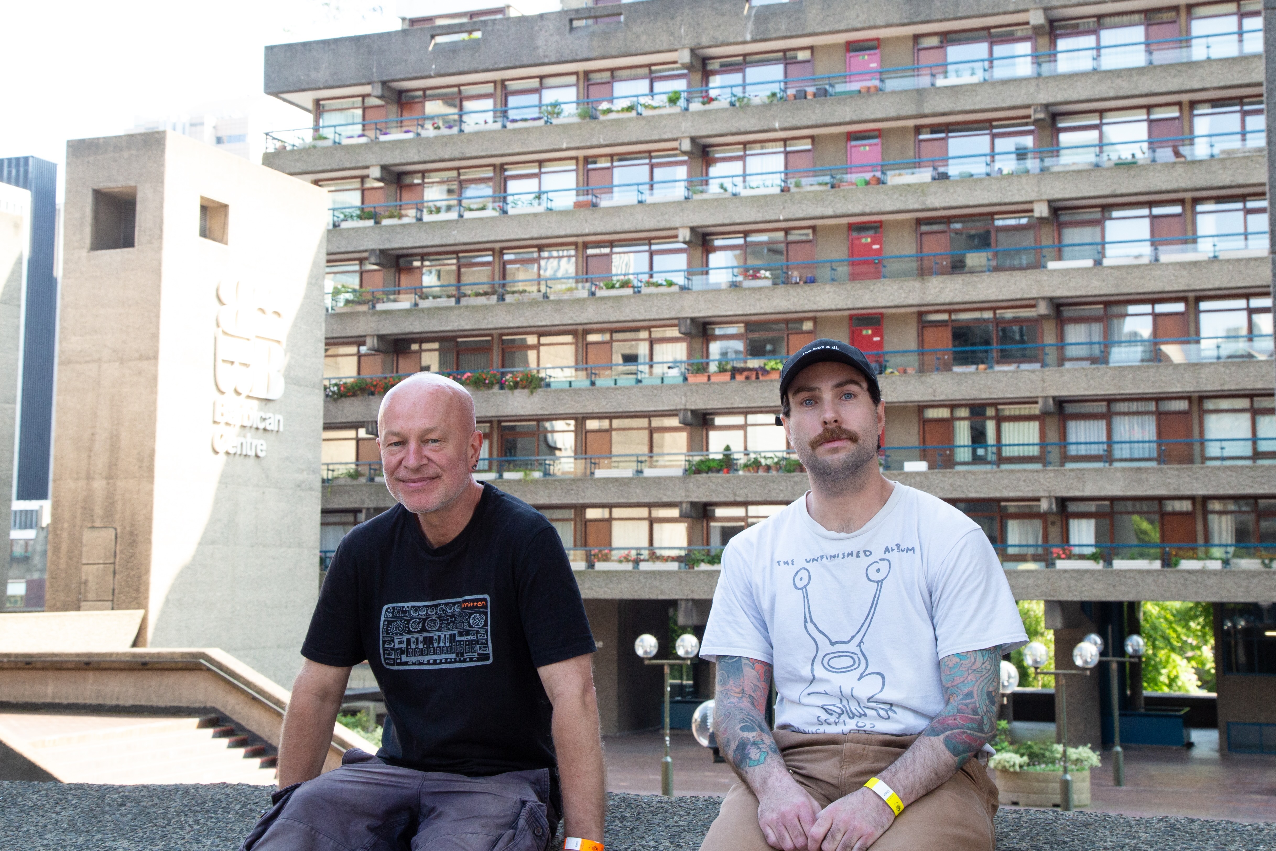 Two men sitting on concrete steps in front of a multi-storey residential building with balconies and coloured awnings.