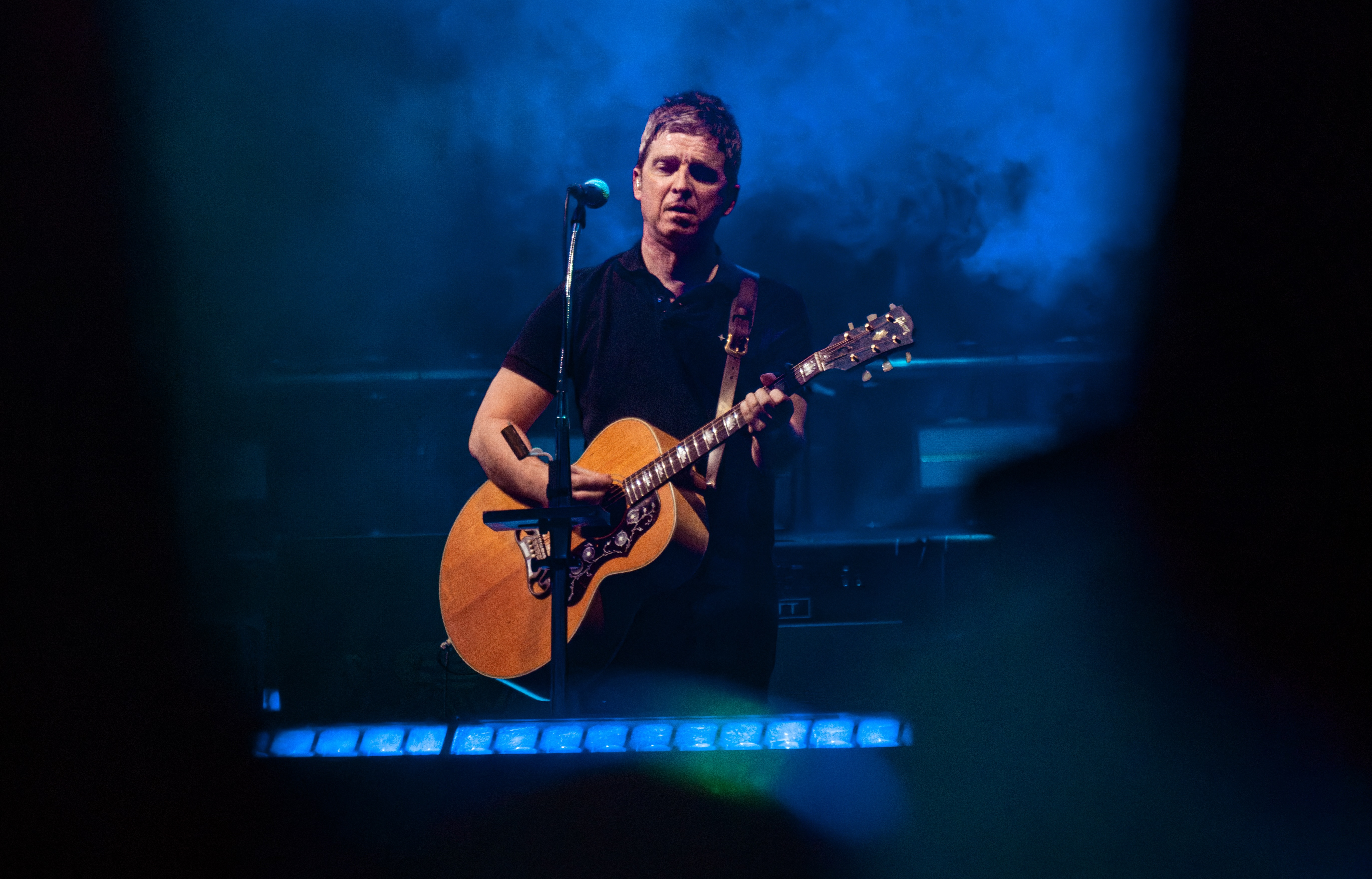 Male musician performing on stage with acoustic guitar and microphone, illuminated by blue stage lighting.