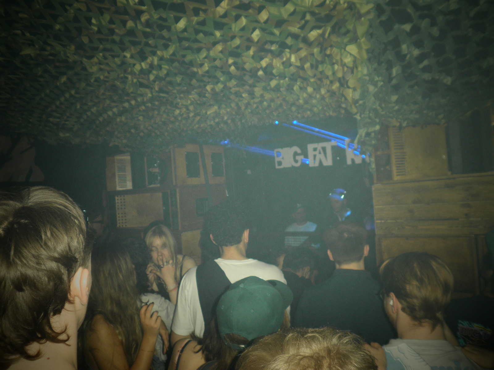 Crowded indoor space with people gathered under green foliage ceiling, blue lighting visible in background, dimly lit atmosphere.