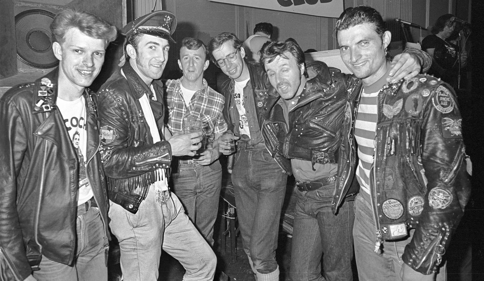 Group of men in leather jackets and casual clothing posing together indoors, black and white photograph with vintage appearance.