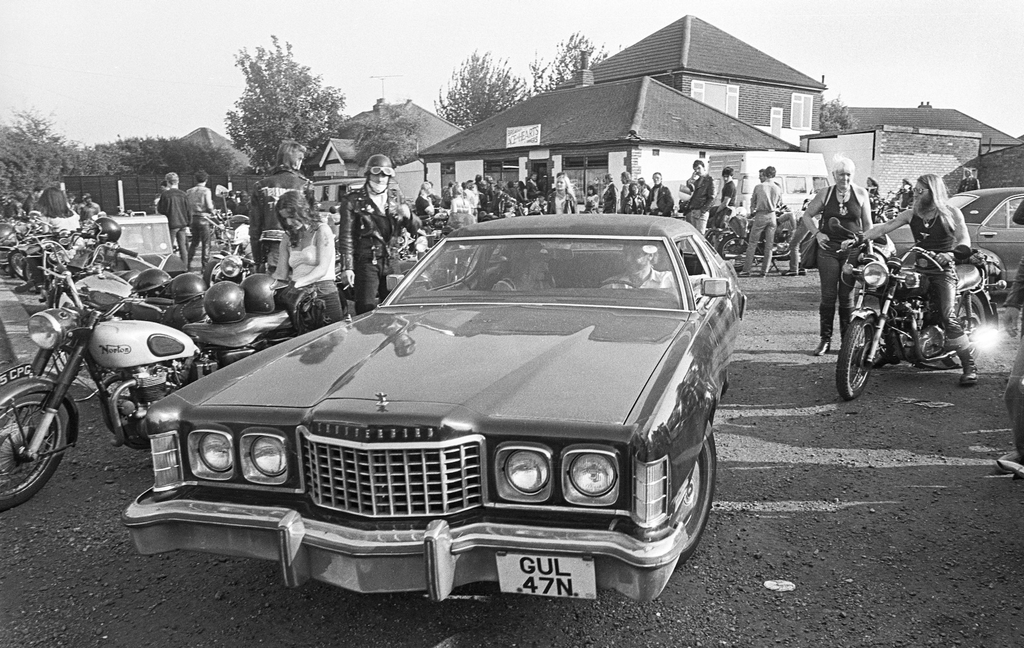 Black and white image showing vintage American car with UK number plate surrounded by motorcycles and crowds at outdoor gathering.