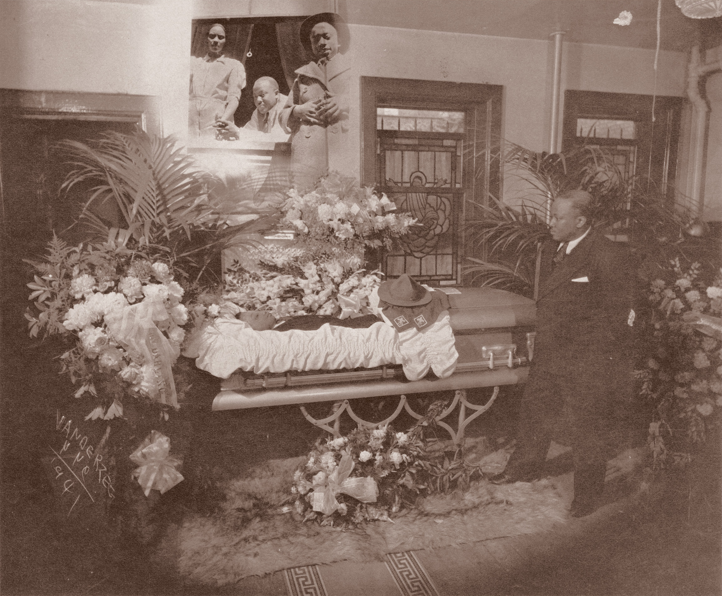 Vintage sepia photograph showing ornate funeral parlour with open casket surrounded by numerous floral arrangements and palm fronds.