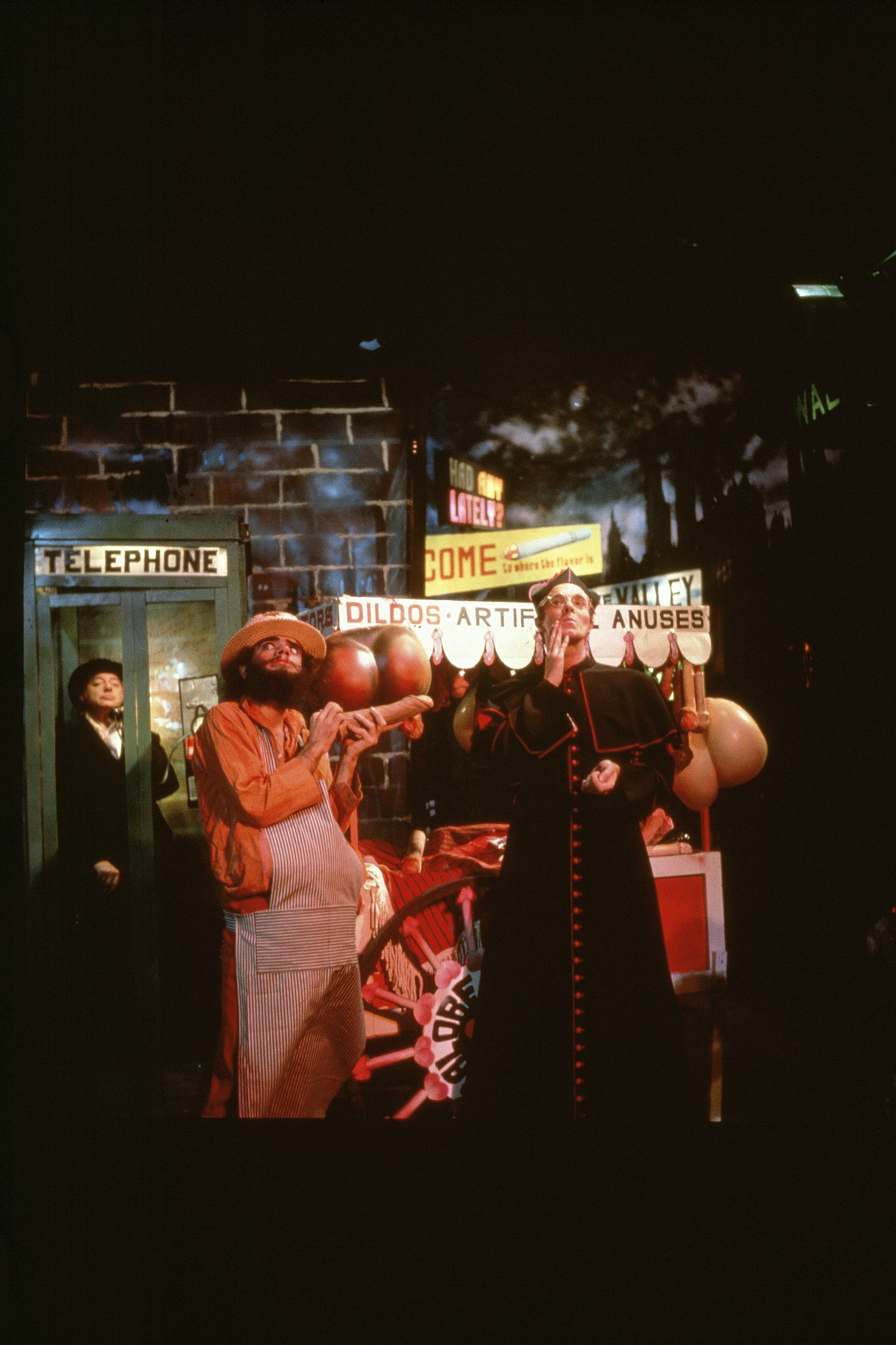Neon-lit street scene with carnival-style attractions and costumed individuals, including a phone booth and signs advertising amusements.