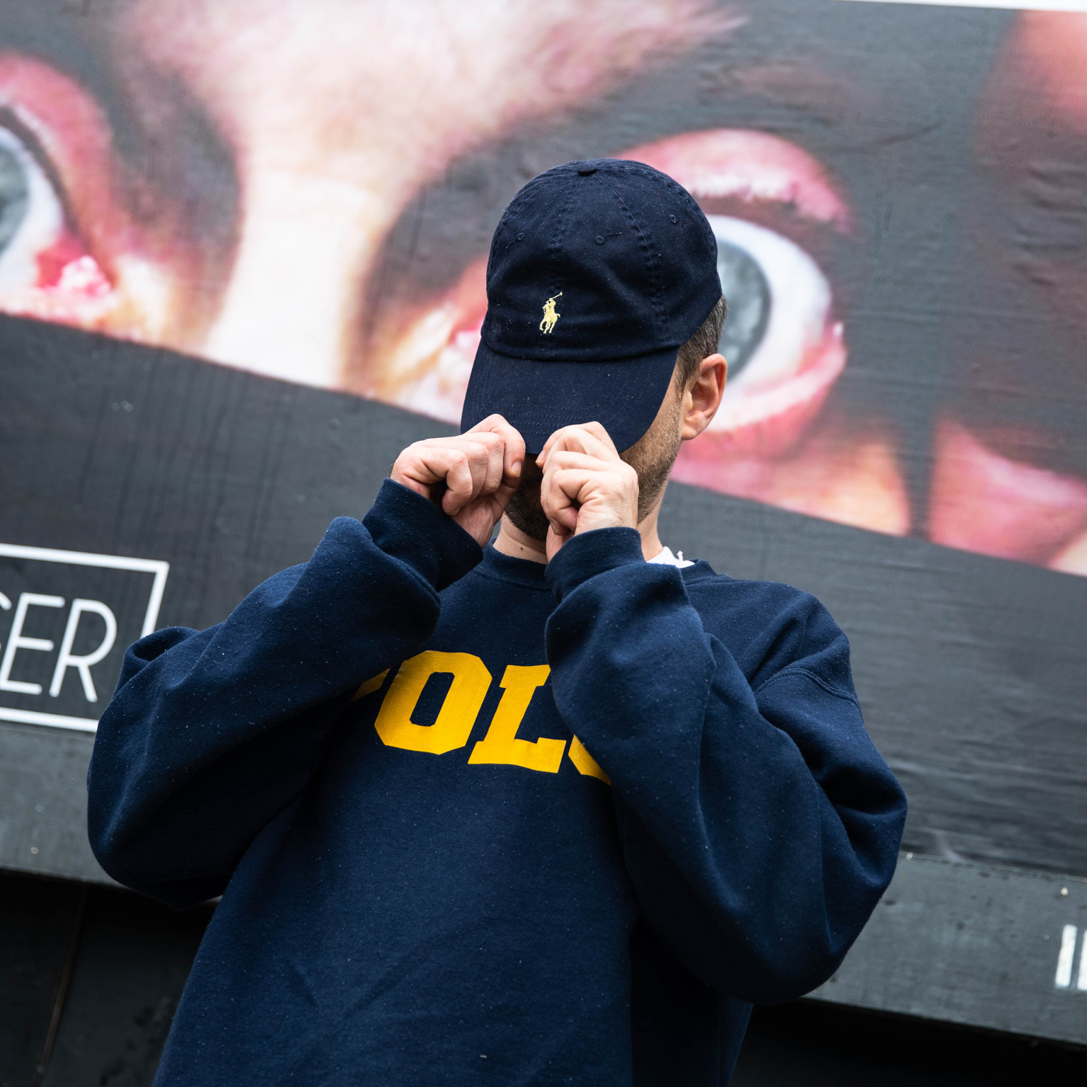 Man in navy hoodie with yellow text and black cap adjusting hood, large screen with close-up eyes visible in background.