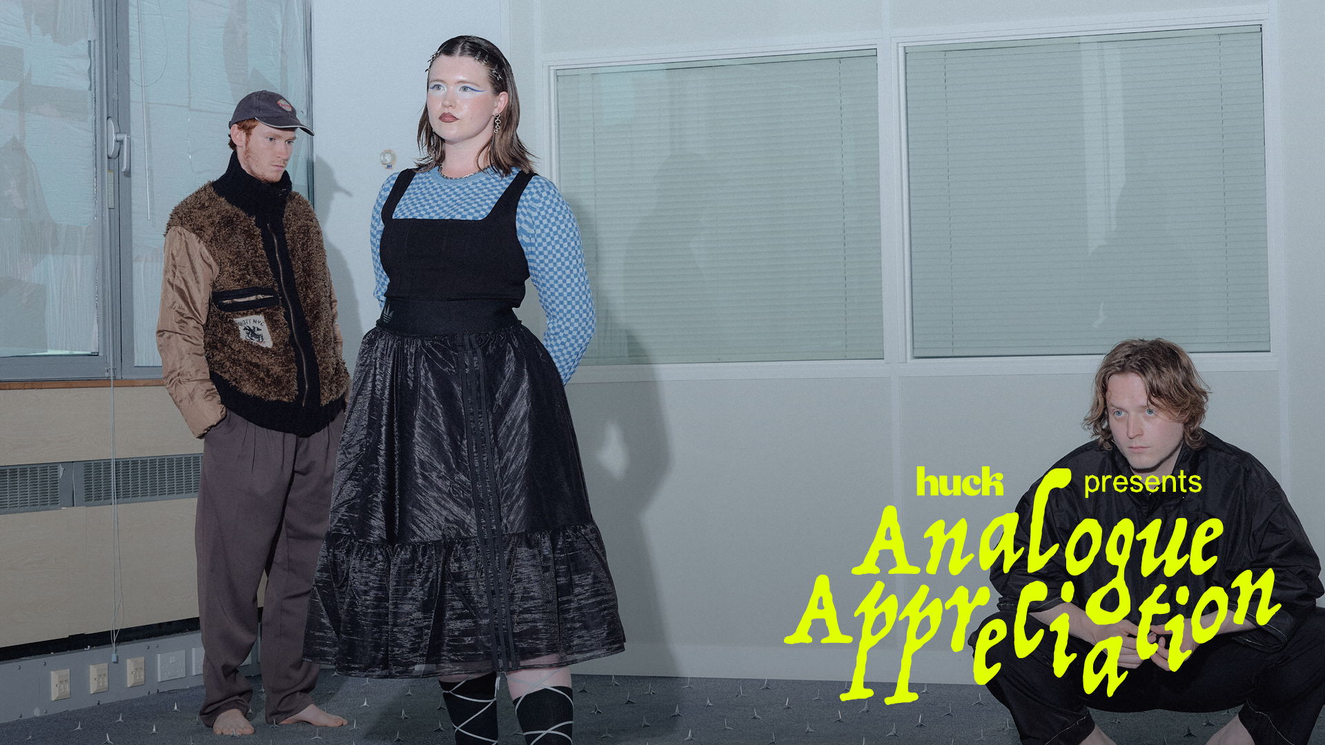 Three people in white-walled room with windows. Woman centre wears black pinafore dress over blue top. Yellow text reads "Analogue Appreciation" bottom right.