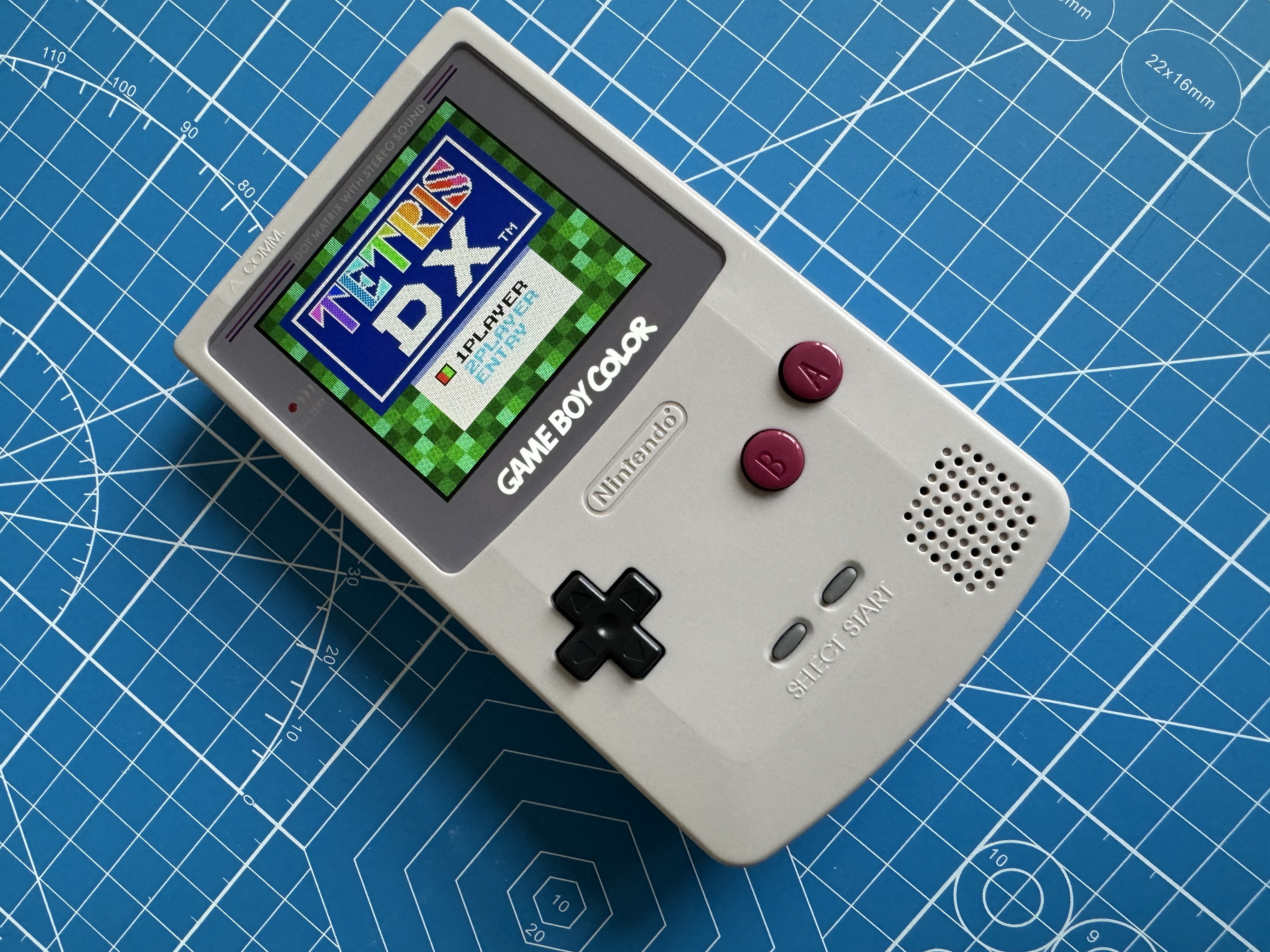 Retro-style handheld game console with colourful screen and buttons on a blue patterned surface.
