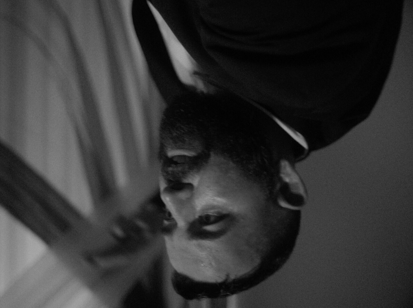 Black and white image of person's face upside down, mouth slightly open, dark hair visible, dramatic lighting with shadows.