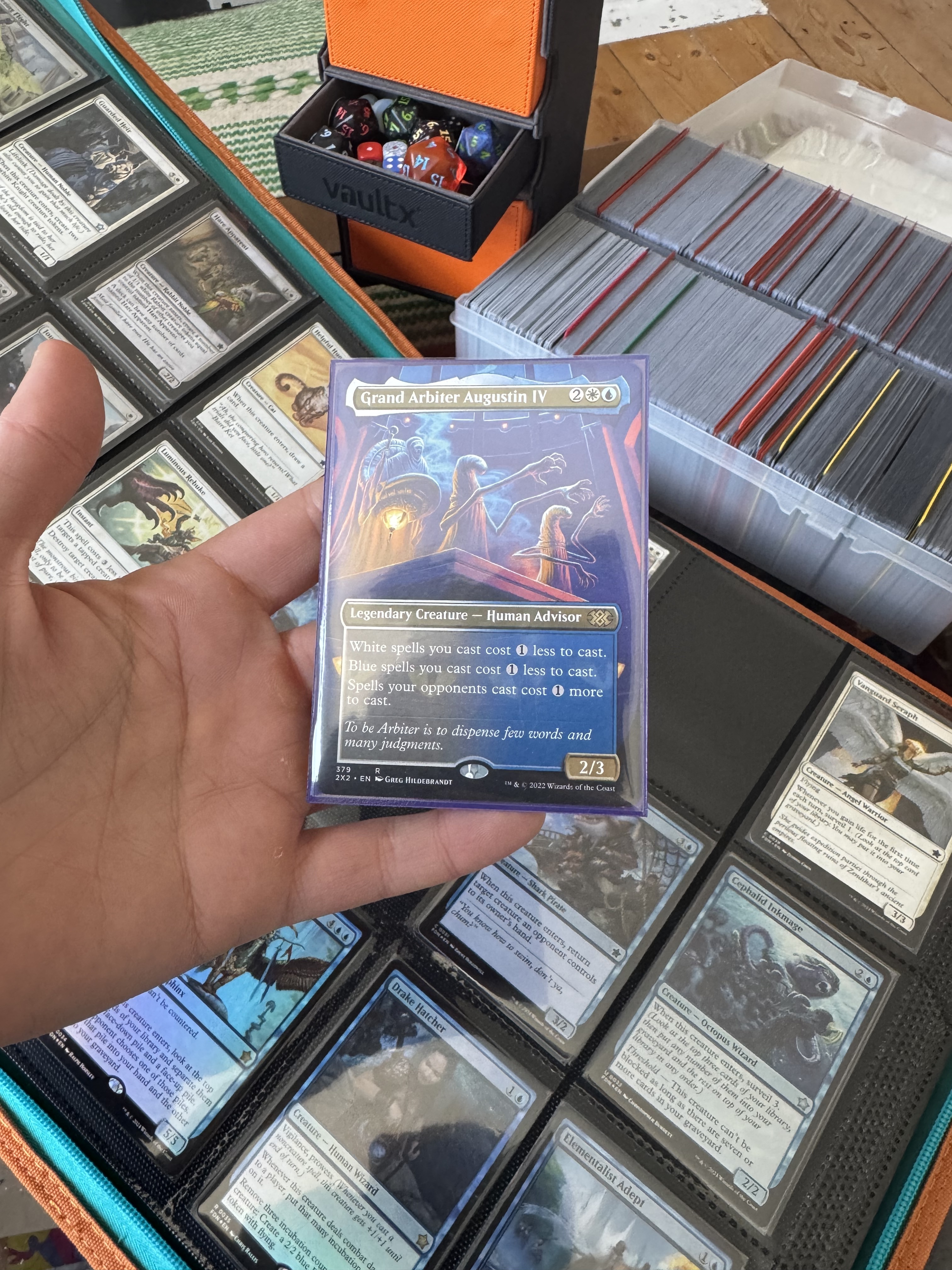 Hand holding a Magic: The Gathering trading card surrounded by piles of other cards, with a cardboard storage box in the background.