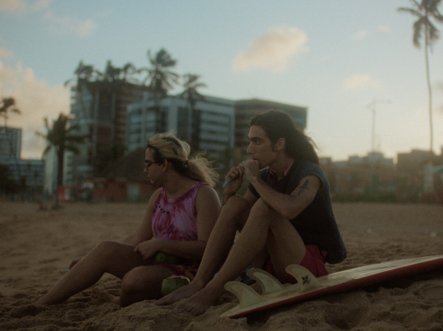 Two people sitting on beach sand beside surfboard, with palm trees and modern building in background under cloudy sky.