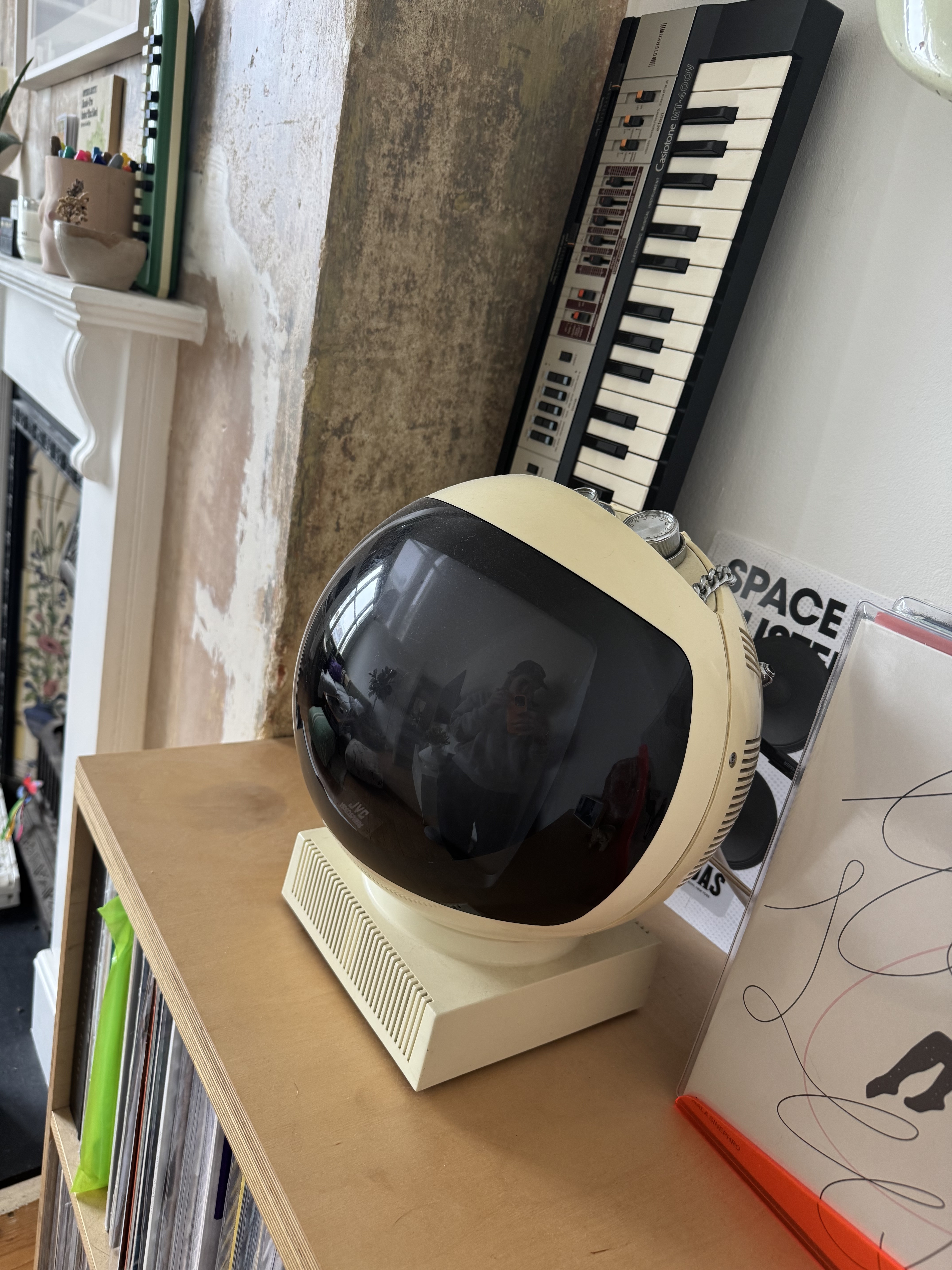 Round cream-coloured audio speaker with black curved display, placed on wooden shelf next to musical keyboard.