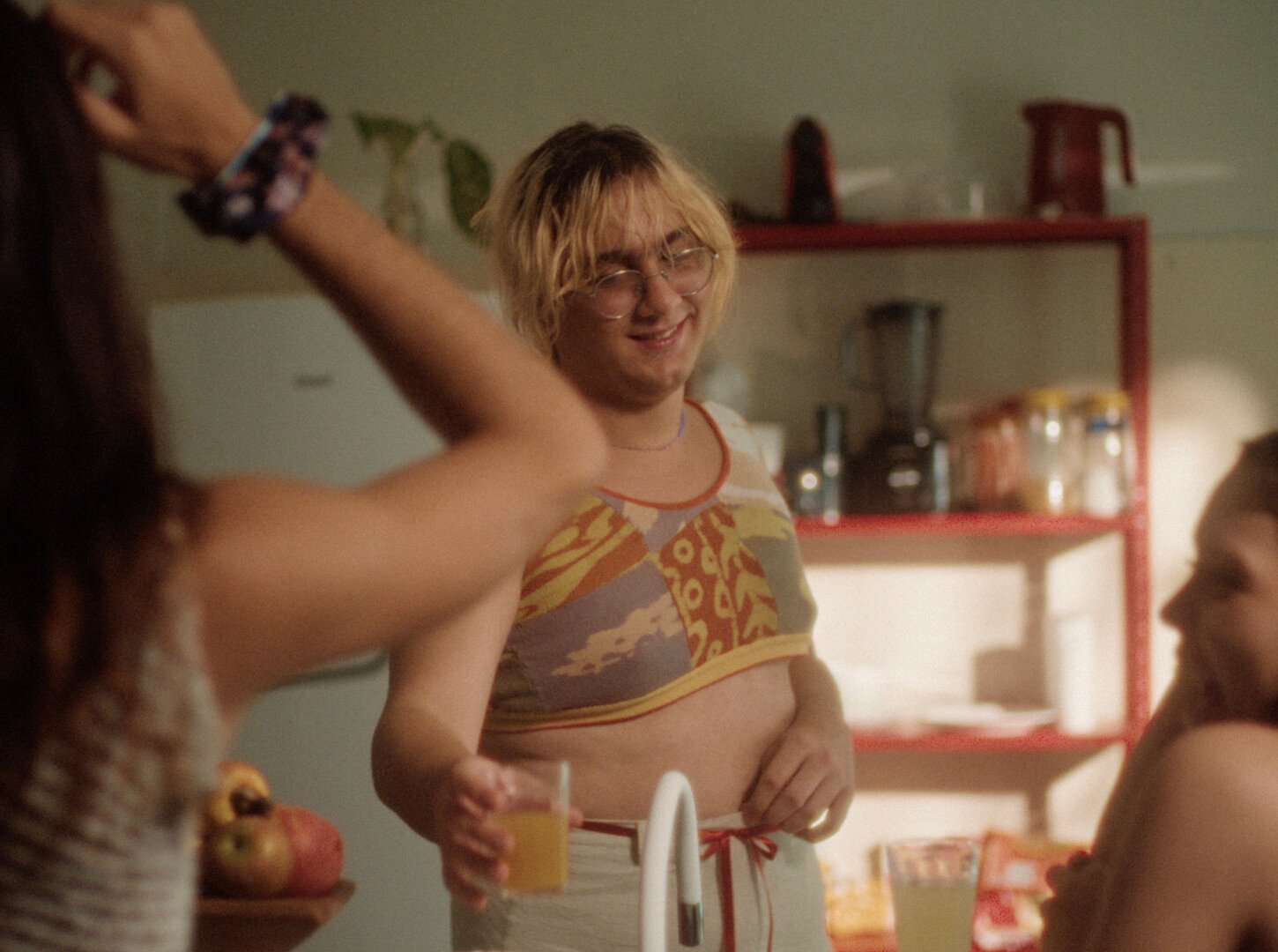 Blonde person in patterned bikini top standing in kitchen with red shelving, arms raised, others partially visible in frame.