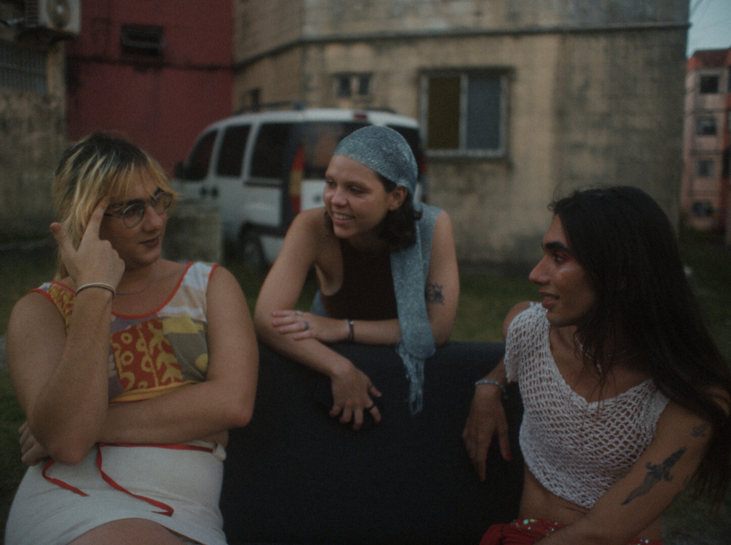 Three women sitting together outdoors, one in floral top, one in blue headscarf and dark clothing, one in white mesh top.