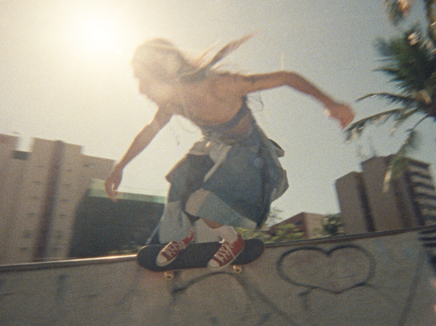 Person skateboarding on concrete surface with palm trees and buildings in background, bright sunlight creating lens flare effect.