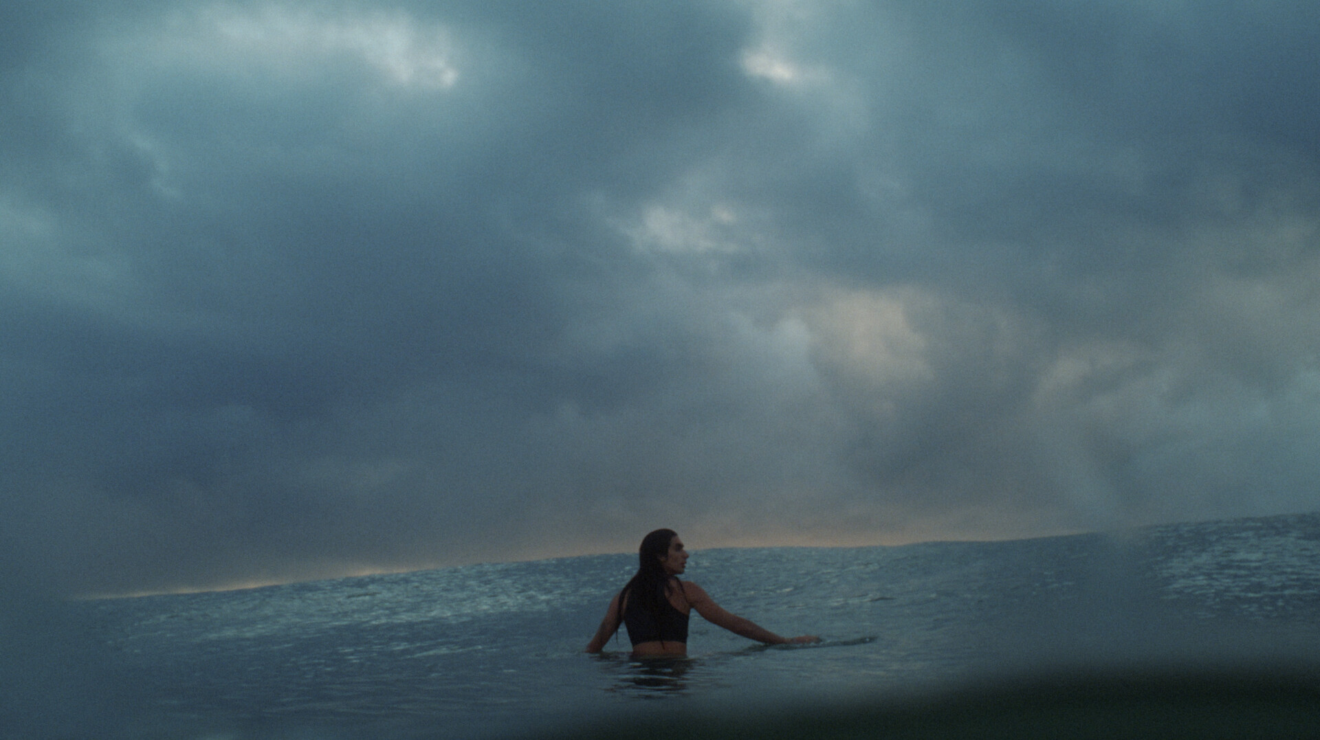 Person swimming in choppy blue-green ocean water under overcast grey sky with storm clouds.