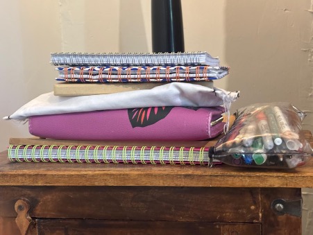 Stack of wrapped notebooks and stationery items on wooden table, featuring striped patterns, pink covers, and clear packaging.