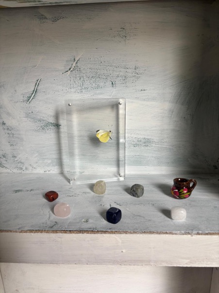 White wooden shelf with clear glass box containing yellow object, surrounded by colourful stones and small decorative bowl.