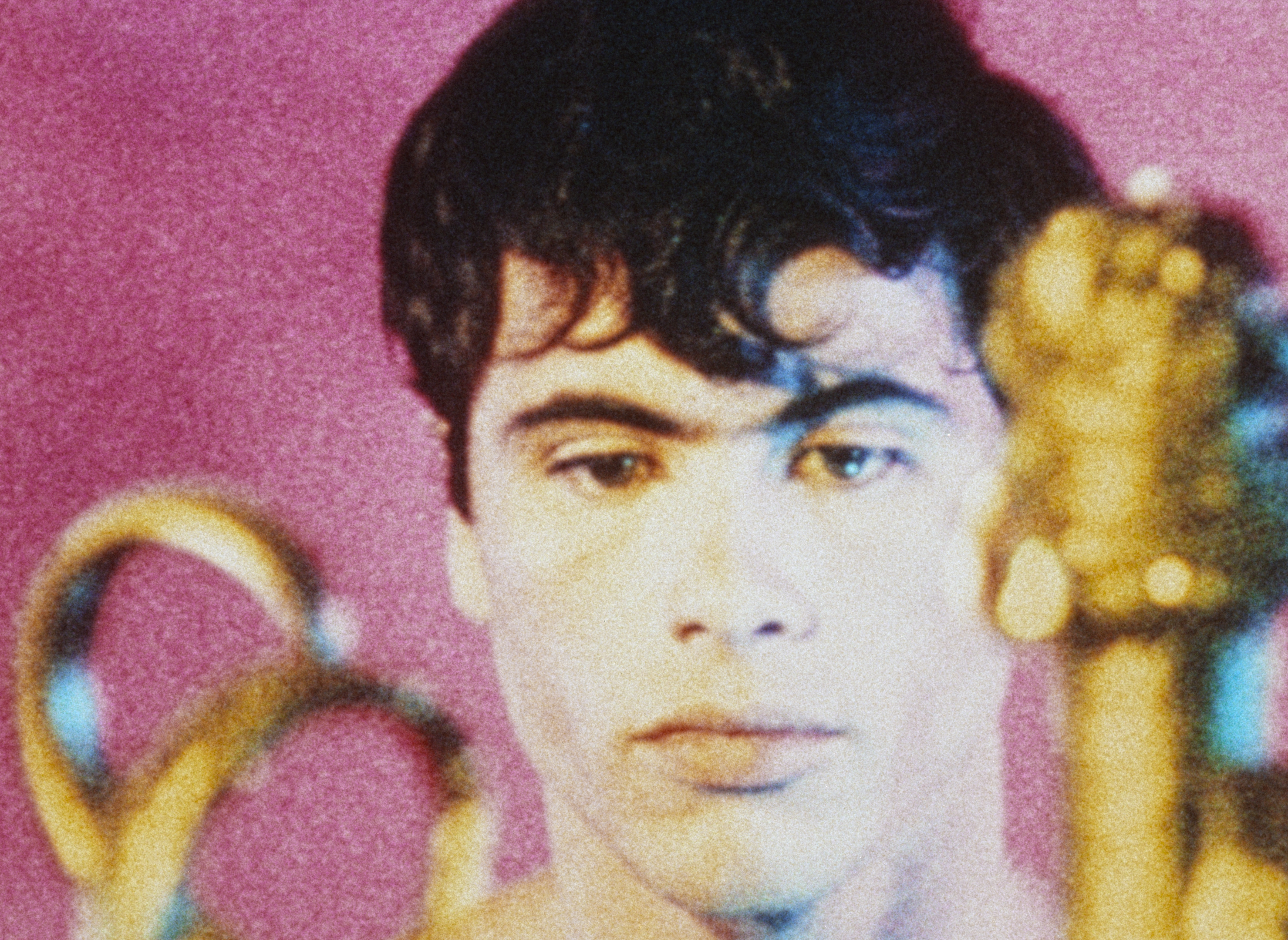 A close-up portrait of a young man with dark hair and intense eyes against a vibrant pink background.