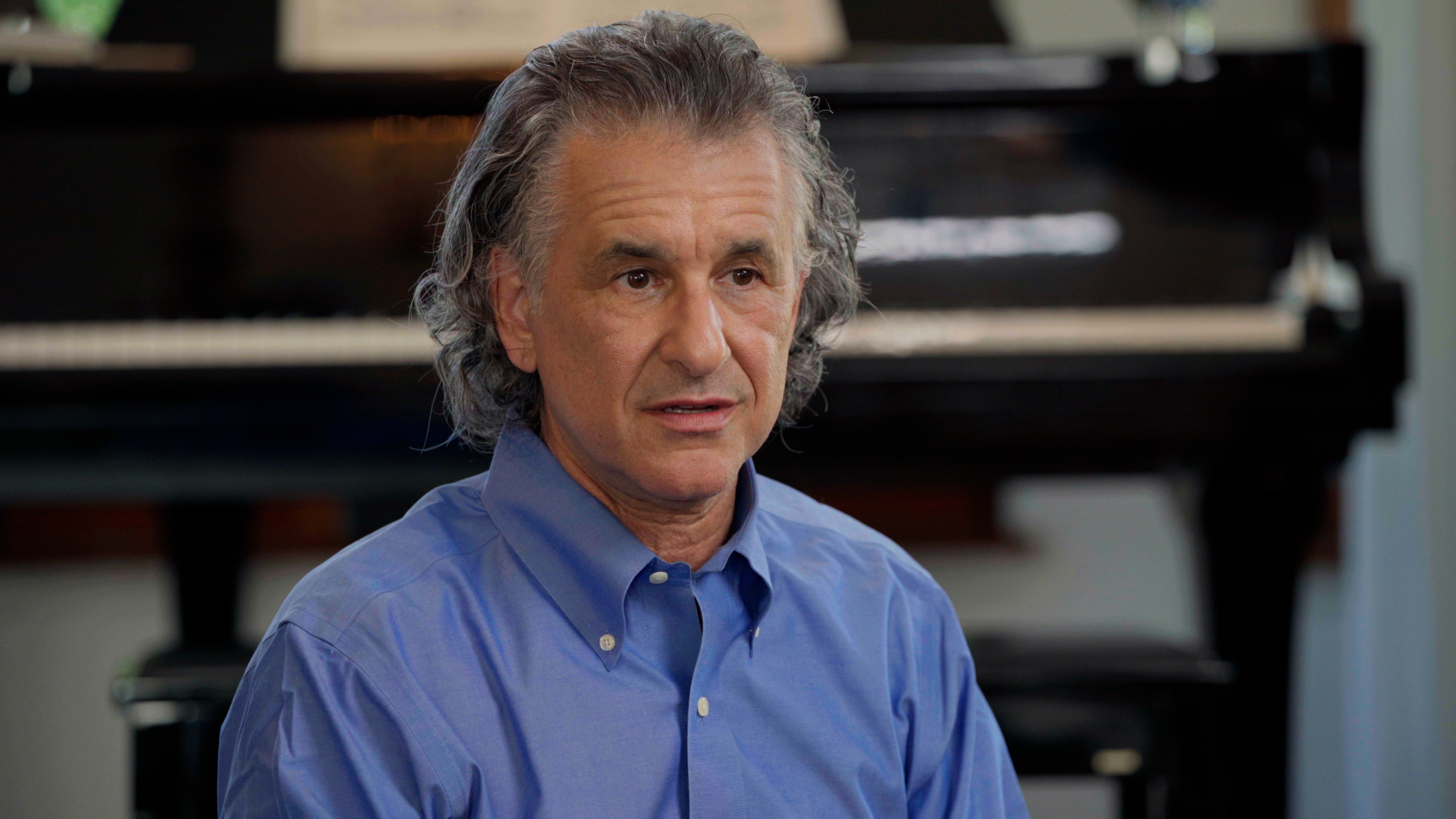 Headshot of an older man with greying curly hair wearing a blue collared shirt.