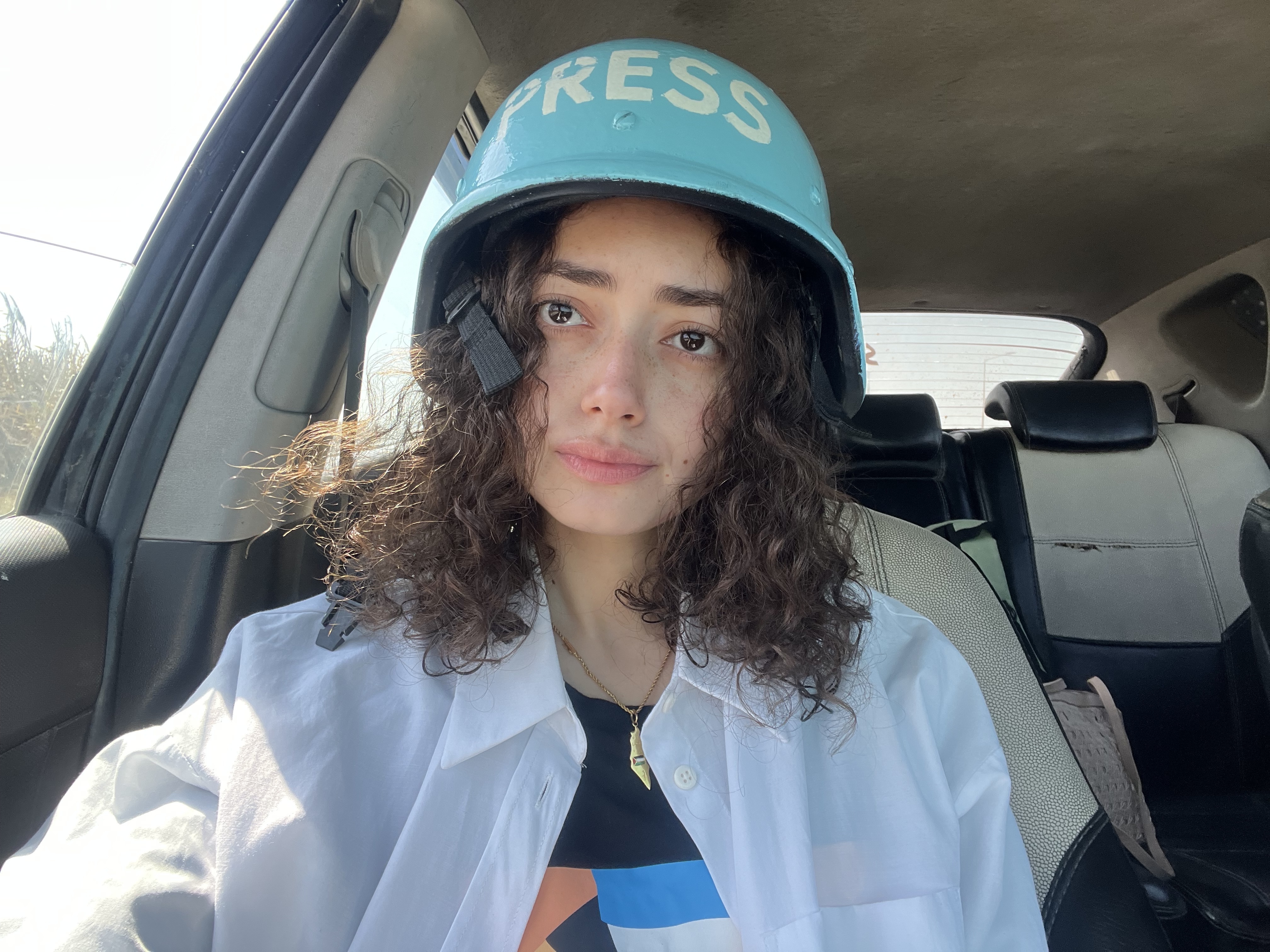 A person wearing a turquoise "Press" cap and a white jacket, with curly hair visible.