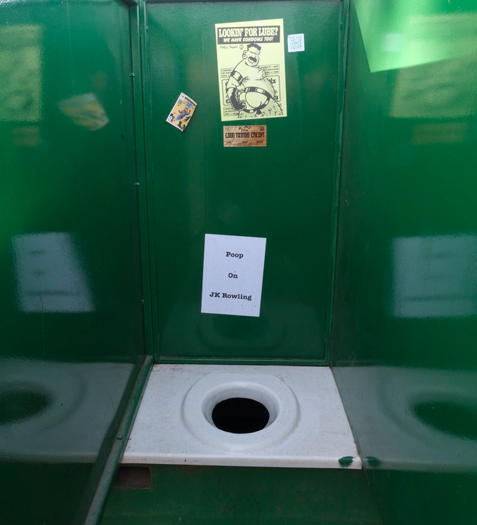 Green portable toilet interior with white seat, posters on back wall, and handwritten sign reading "Poop On JK Rowling"