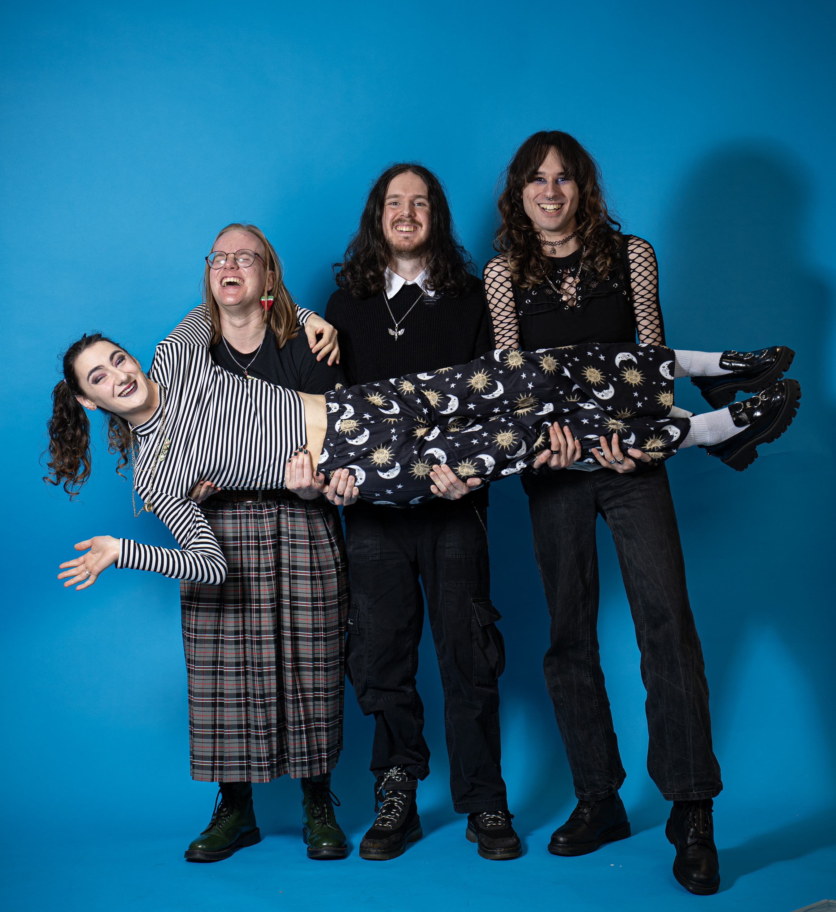 Four people against blue backdrop, three holding one person horizontally. Mixed clothing: striped top, plaid skirt, dark outfits, boots.