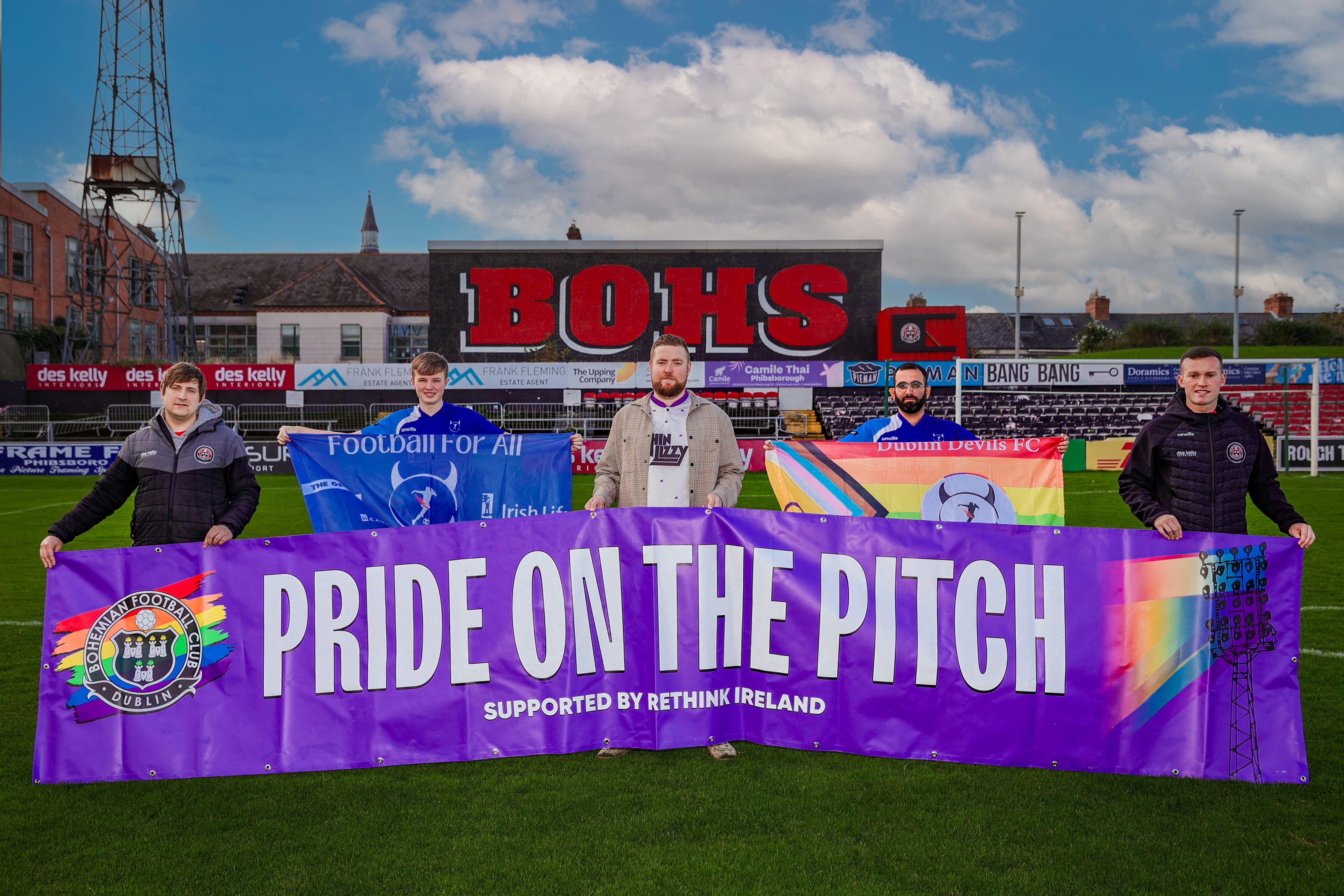 Supporters holding a Pride on the Pitch banner on a football pitch, featuring the words "BOYS" on a stadium in the background.
