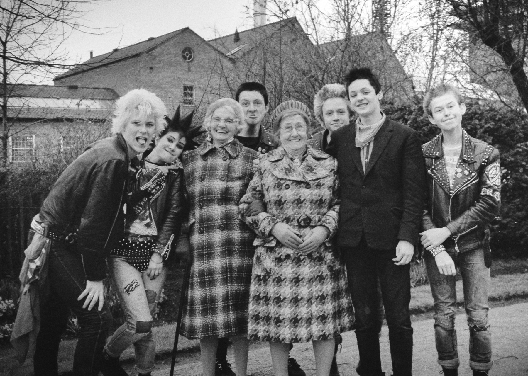 Black and white image showing eight people standing together outdoors, including punk-styled individuals and elderly women in coats, with houses visible in background.