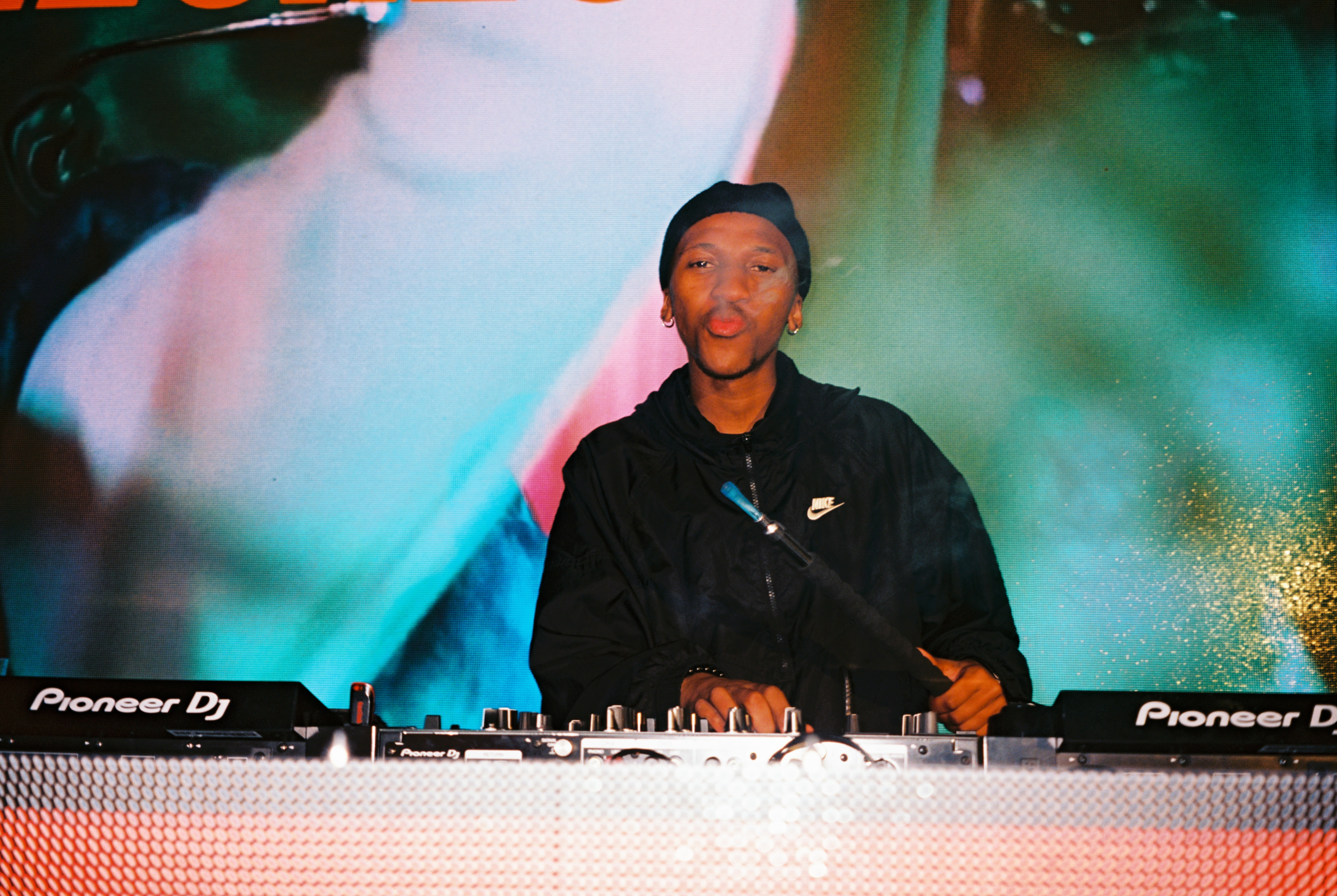 Young DJ wearing black clothing and a cap, playing music on Pioneer equipment in front of psychedelic lighting effects.