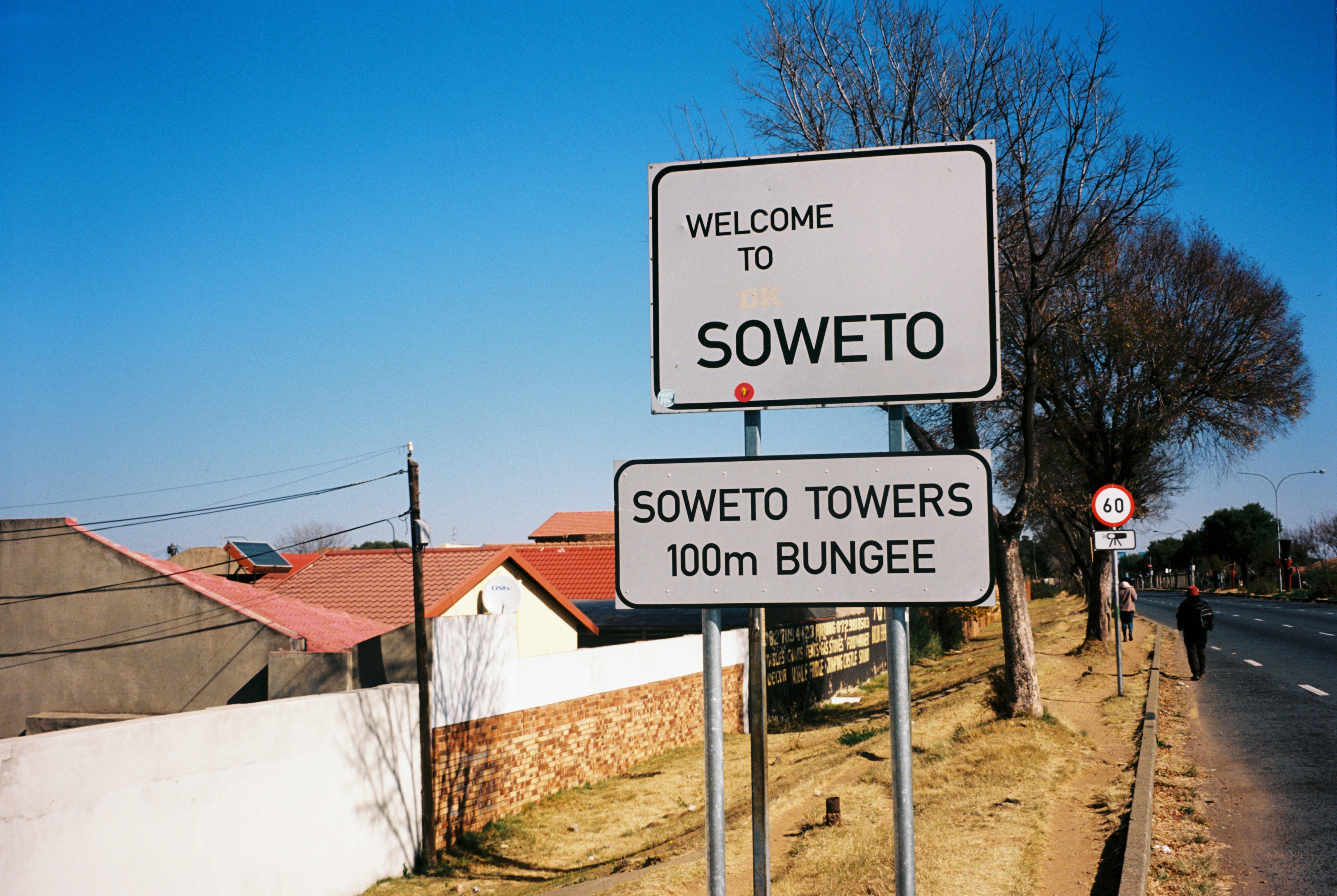 Welcome sign for Soweto, indicating Soweto Towers 100m bungee. Road with buildings and trees visible.