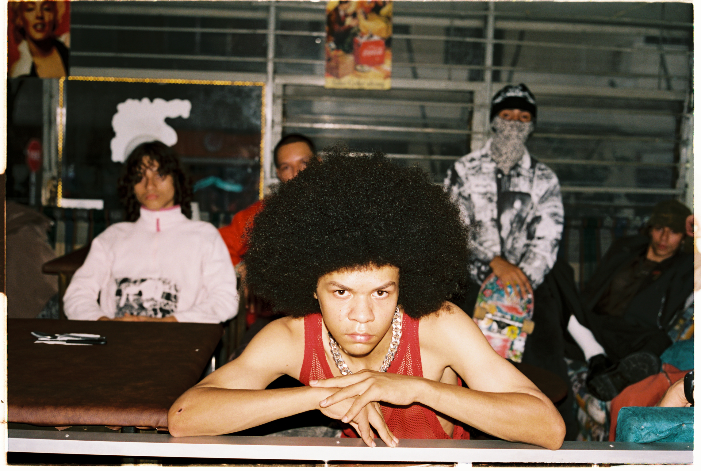 Man with afro hairstyle in red vest leans on counter, arms crossed. Several people visible in background wearing white and patterned clothing.