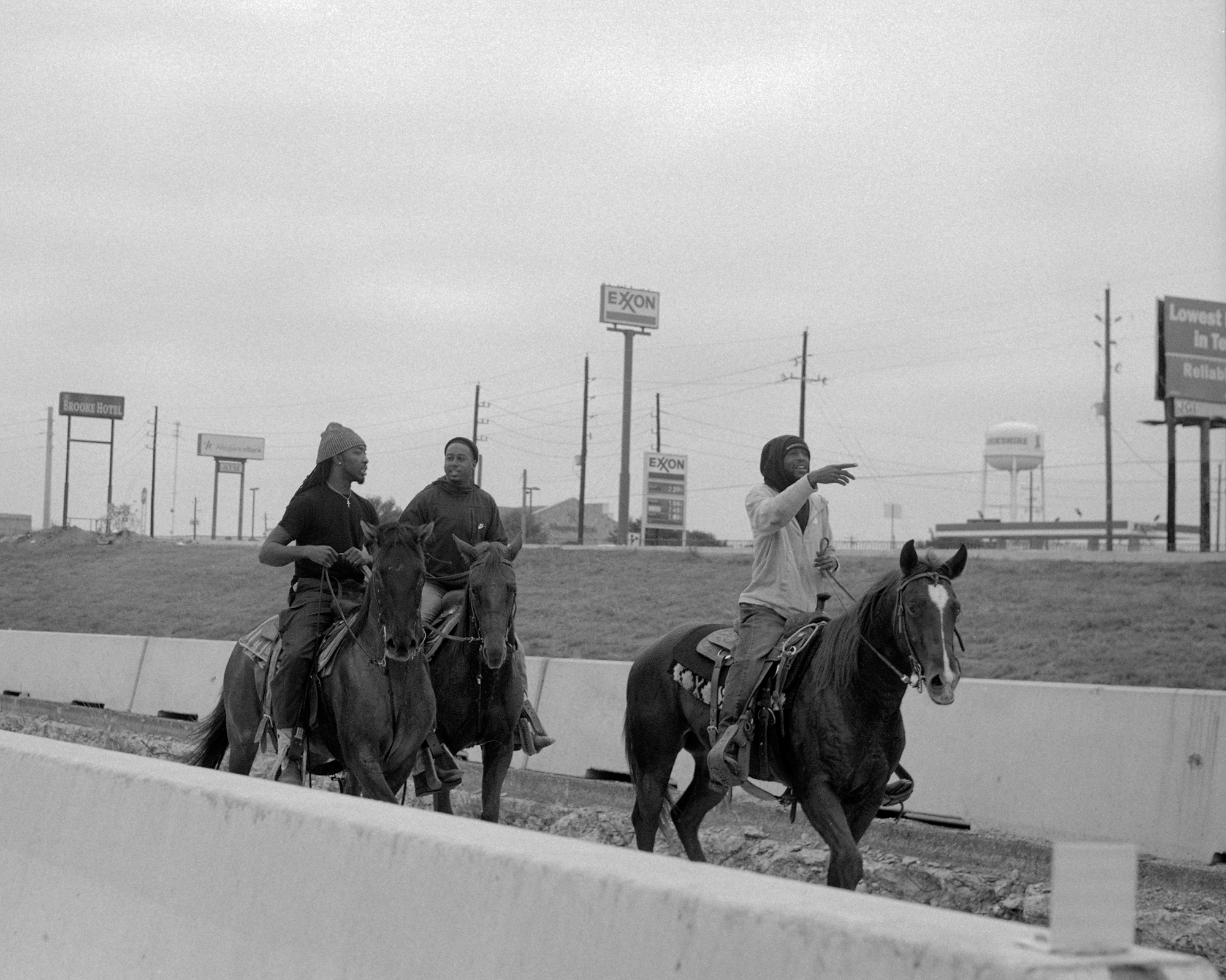 Three people riding horses along a road, with signs and commercial buildings visible in the background.