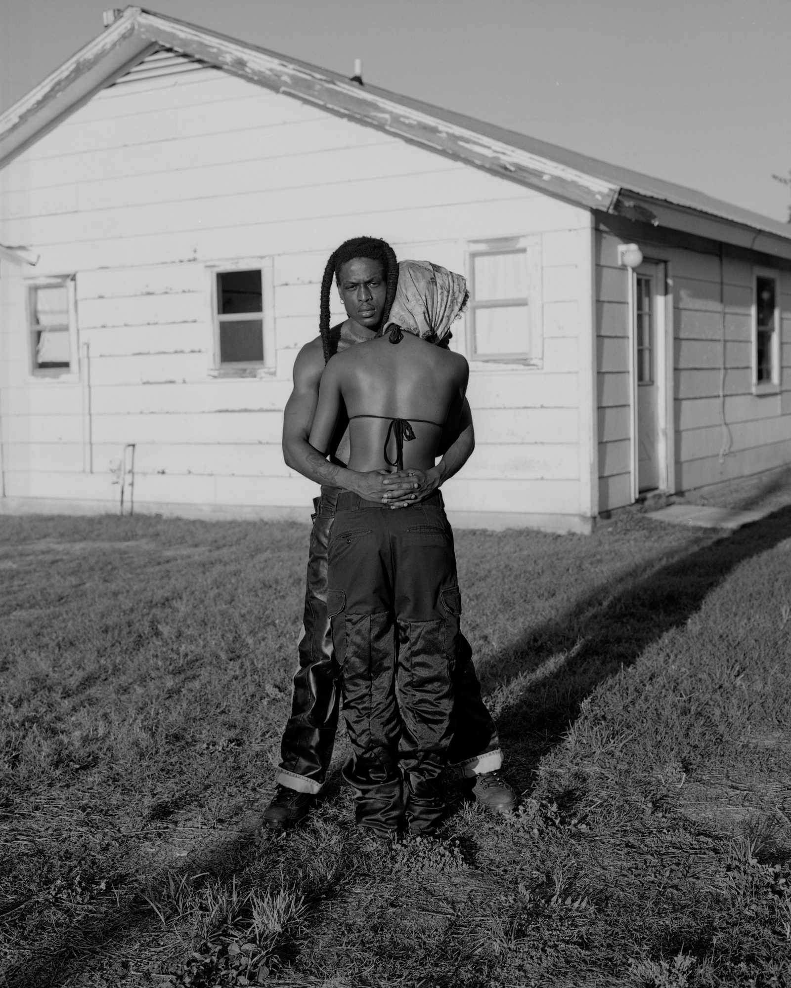 Black and white image of a shirtless man standing in a field in front of a wooden structure.